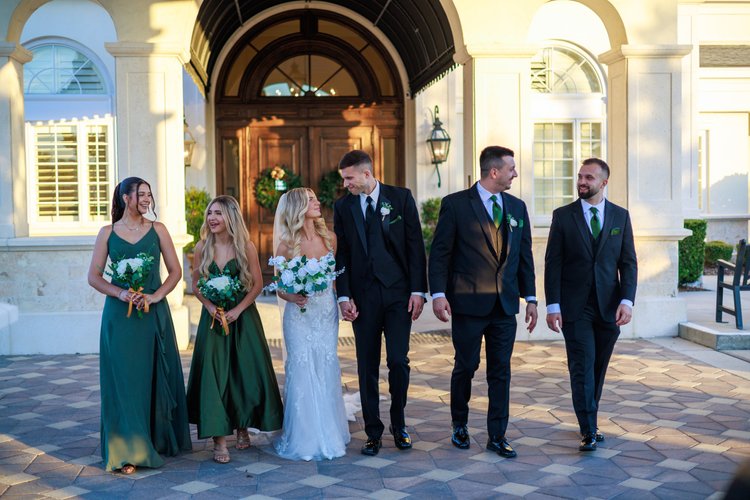 The full wedding party walks together toward the camera outside a white archway entrance, bridesmaids in green and groomsmen in dark suits flanking the couple.