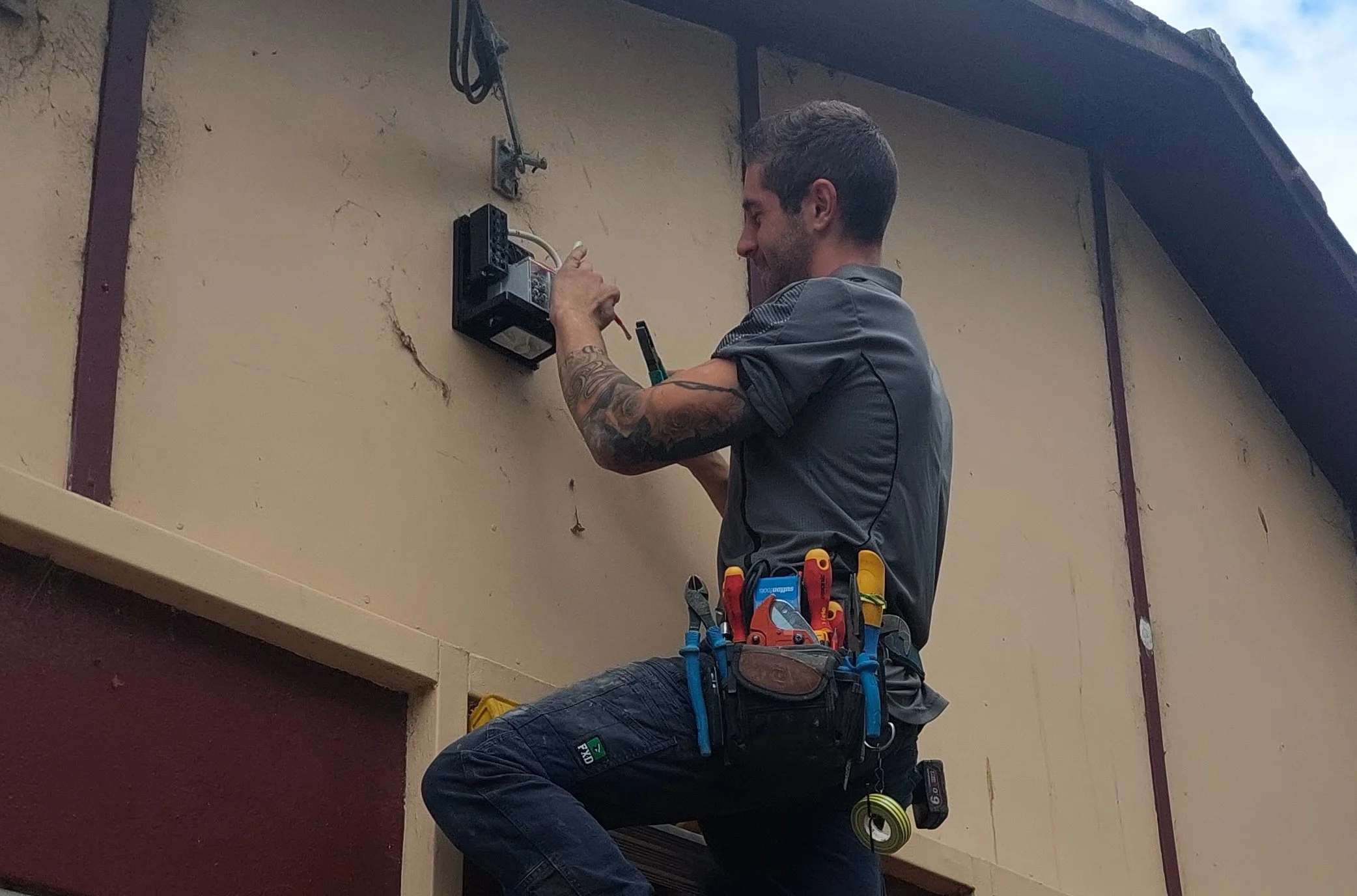 A man with tattoos wearing a gray work shirt and safety tools around his waist installing or repairing an electrical box on a beige wall outside a building.