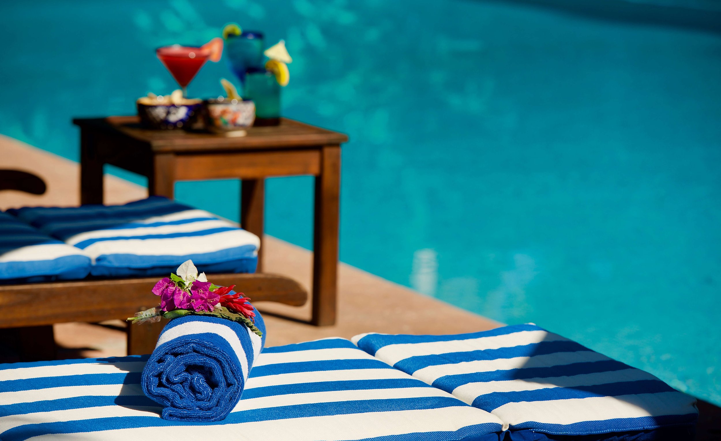 A poolside scene with a blue and white striped lounge chair, a rolled blue towel with pink flowers on top, and a small wooden table holding colorful drinks and bowls of snacks, set against a background of a swimming pool.