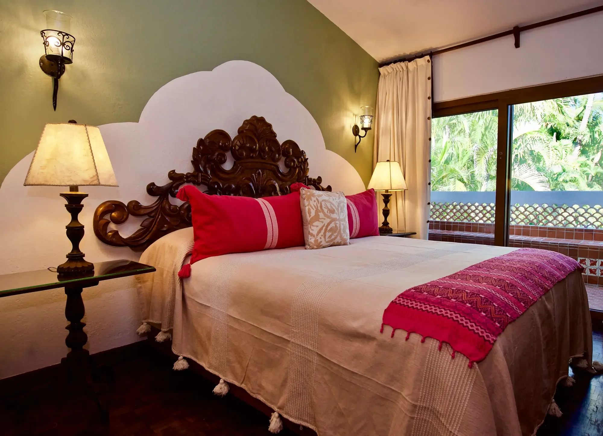 A bedroom with a large wooden carved headboard, red pillows and a beige bedspread. Two lamps provide warm lighting, and a large sliding glass door opens to a balcony with greenery outside.
