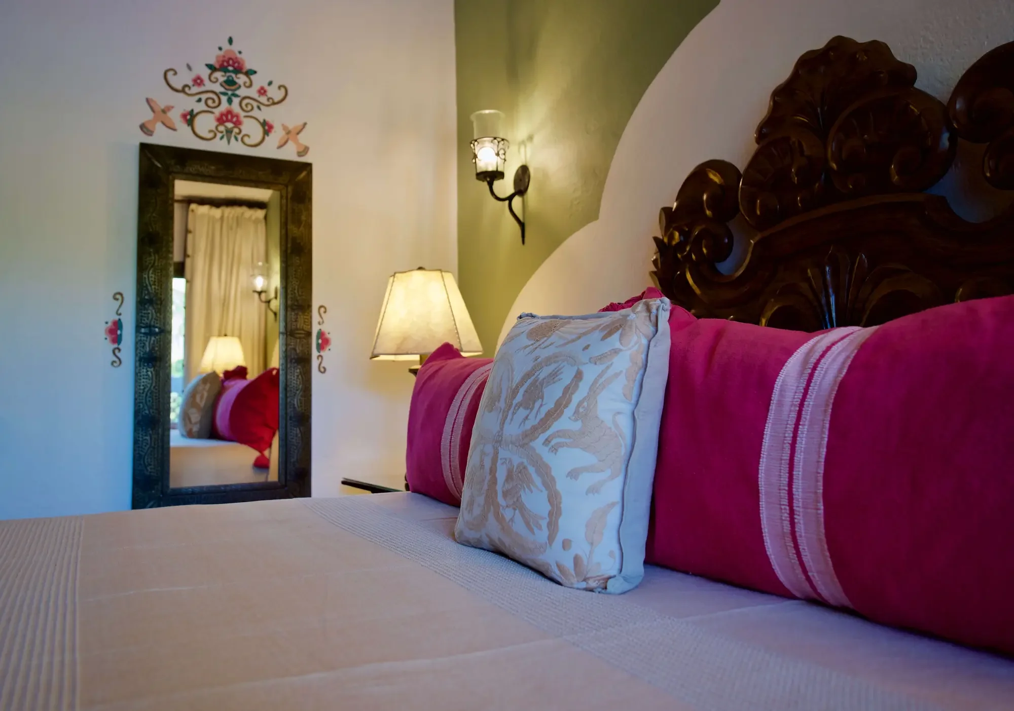 A bedroom with a bed covered with a pink blanket and decorative pillows. A mirror on the wall reflects the opposite side of the room, which has a window with curtains. The room is lit by two wall-mounted lights and a table lamp.