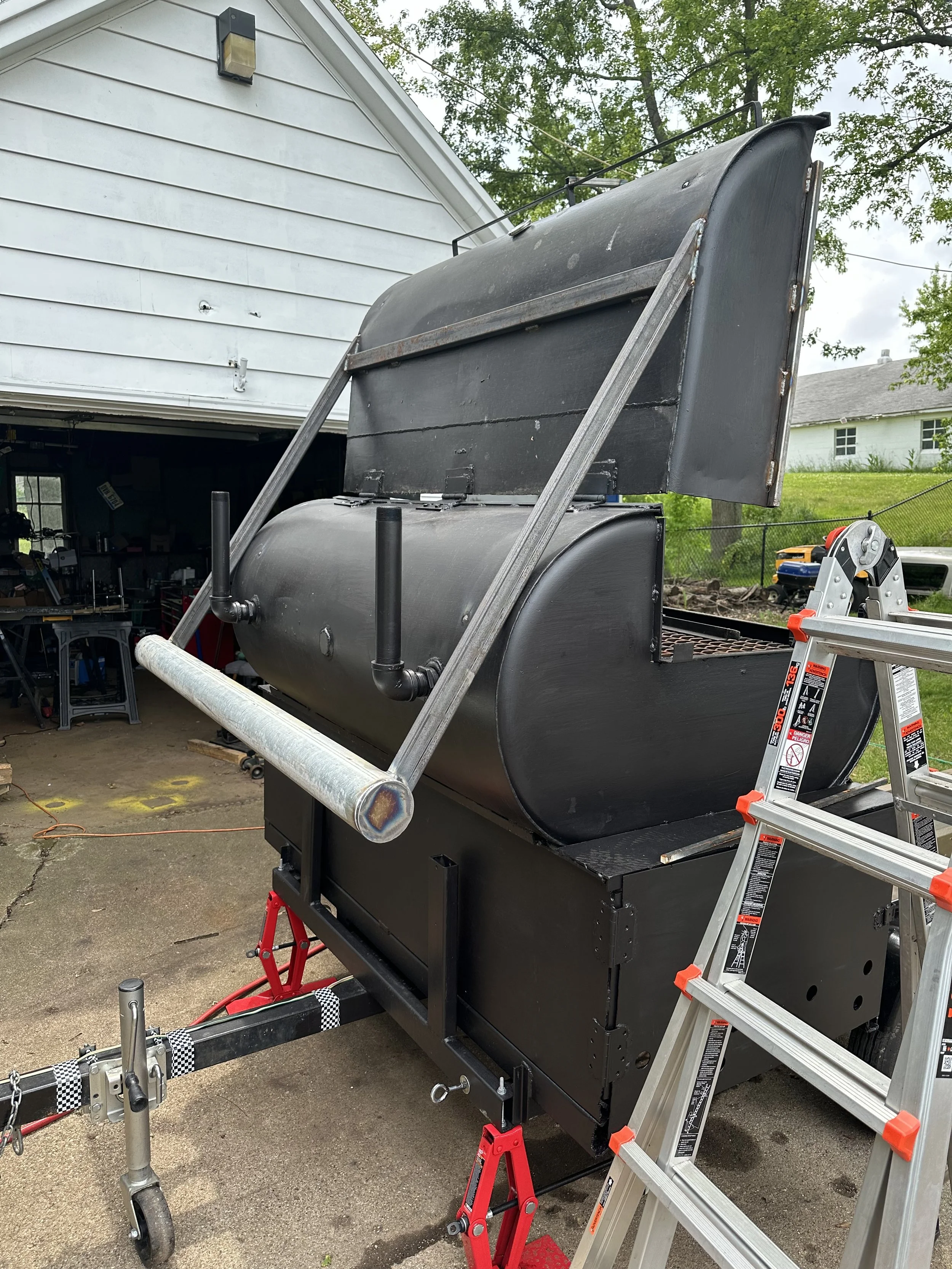 A metal smoker grill with a black finish, on a stand with wheels, is outdoors in a driveway next to a ladder. The lid of the smoker is open, revealing the cooking chamber. The background shows a house, trees, and part of the garage interior.