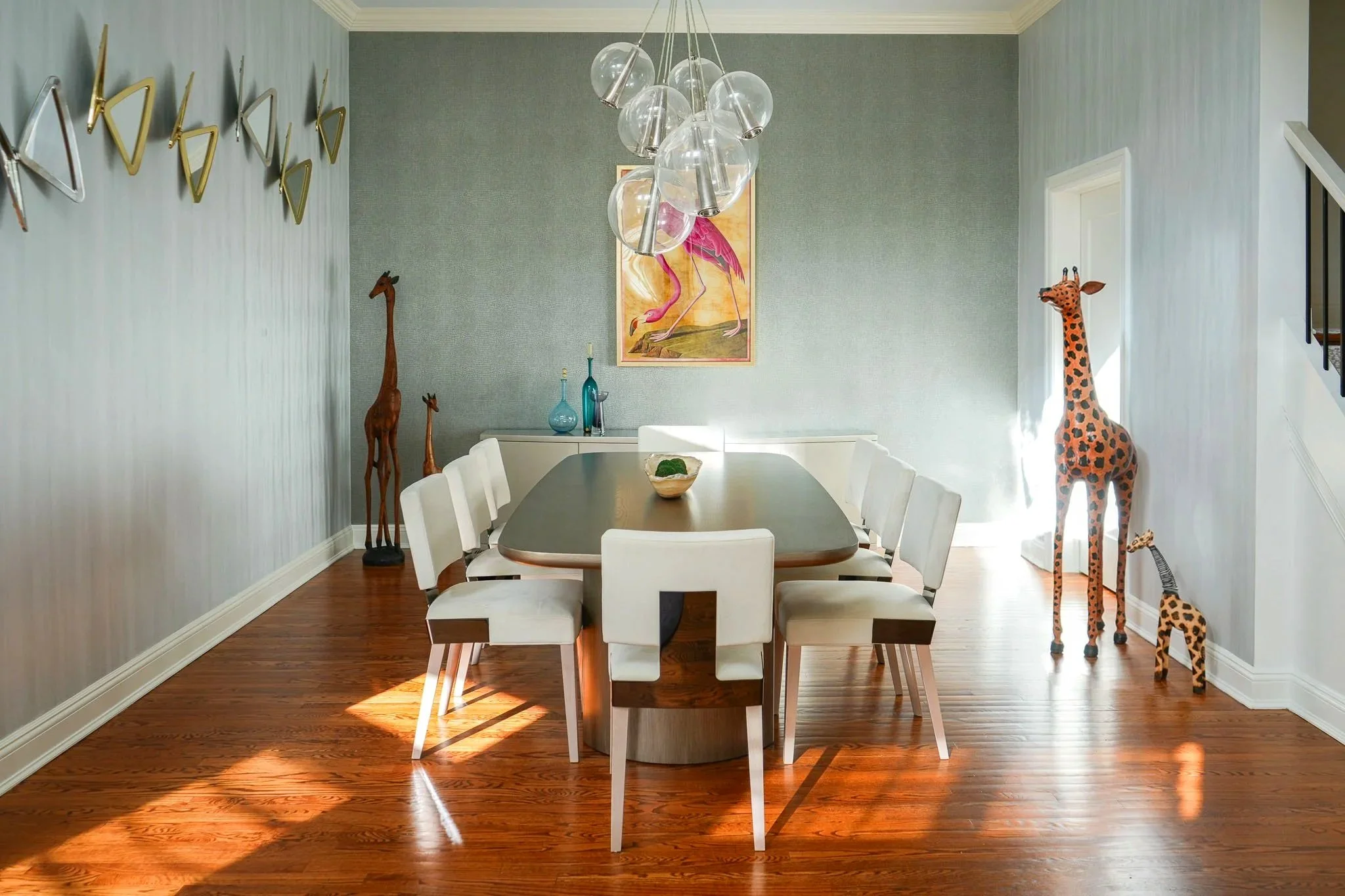 Dining room with a wooden table and eight white chairs, decorated with giraffe and flamingo wall art, and a modern chandelier hanging from the ceiling.