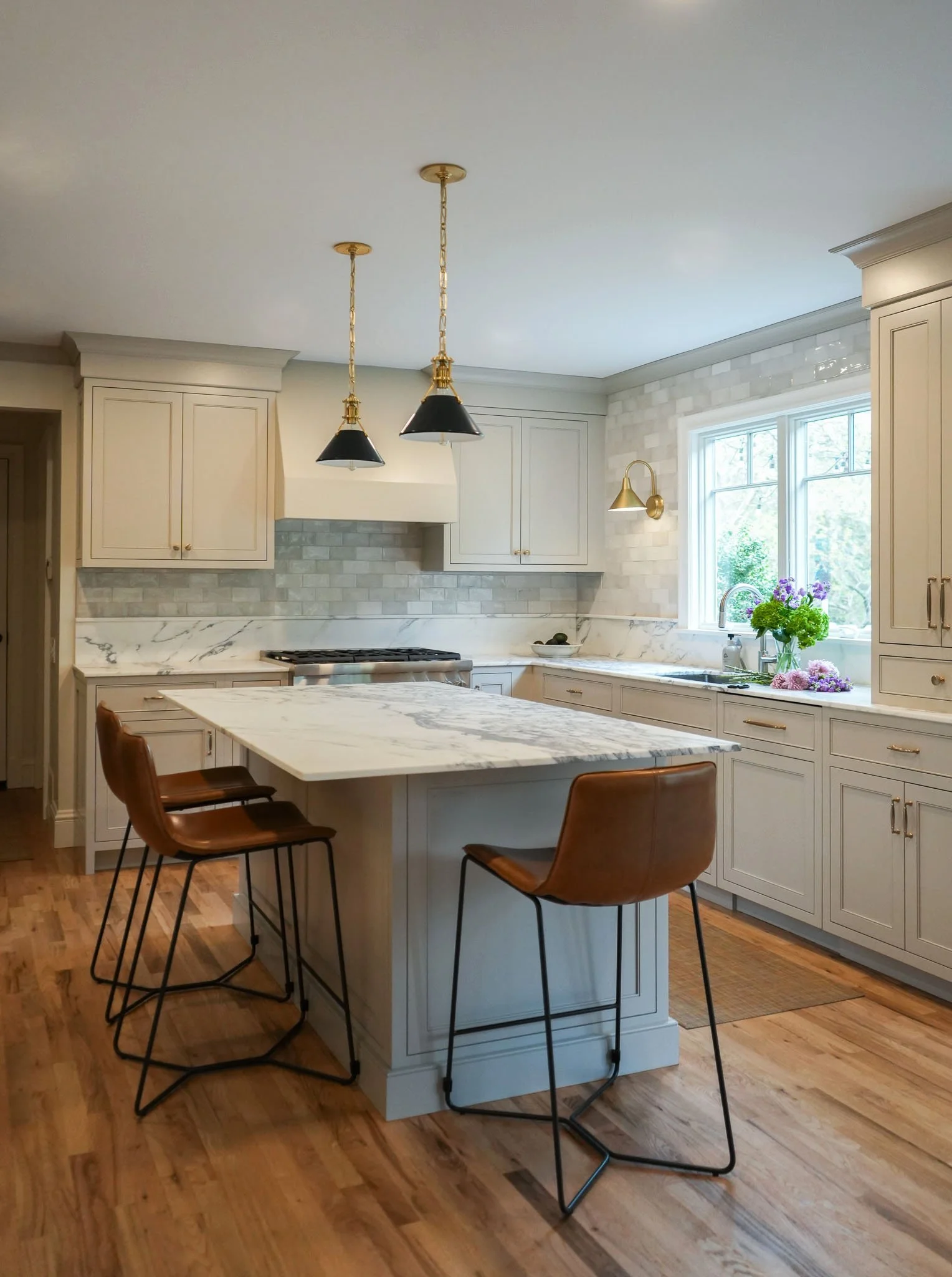 A modern kitchen with white cabinetry, a marble island, and a wooden floor. Two black and gold pendant lights hang above the island. A window with greenery outside and a vase of purple flowers on the counter.