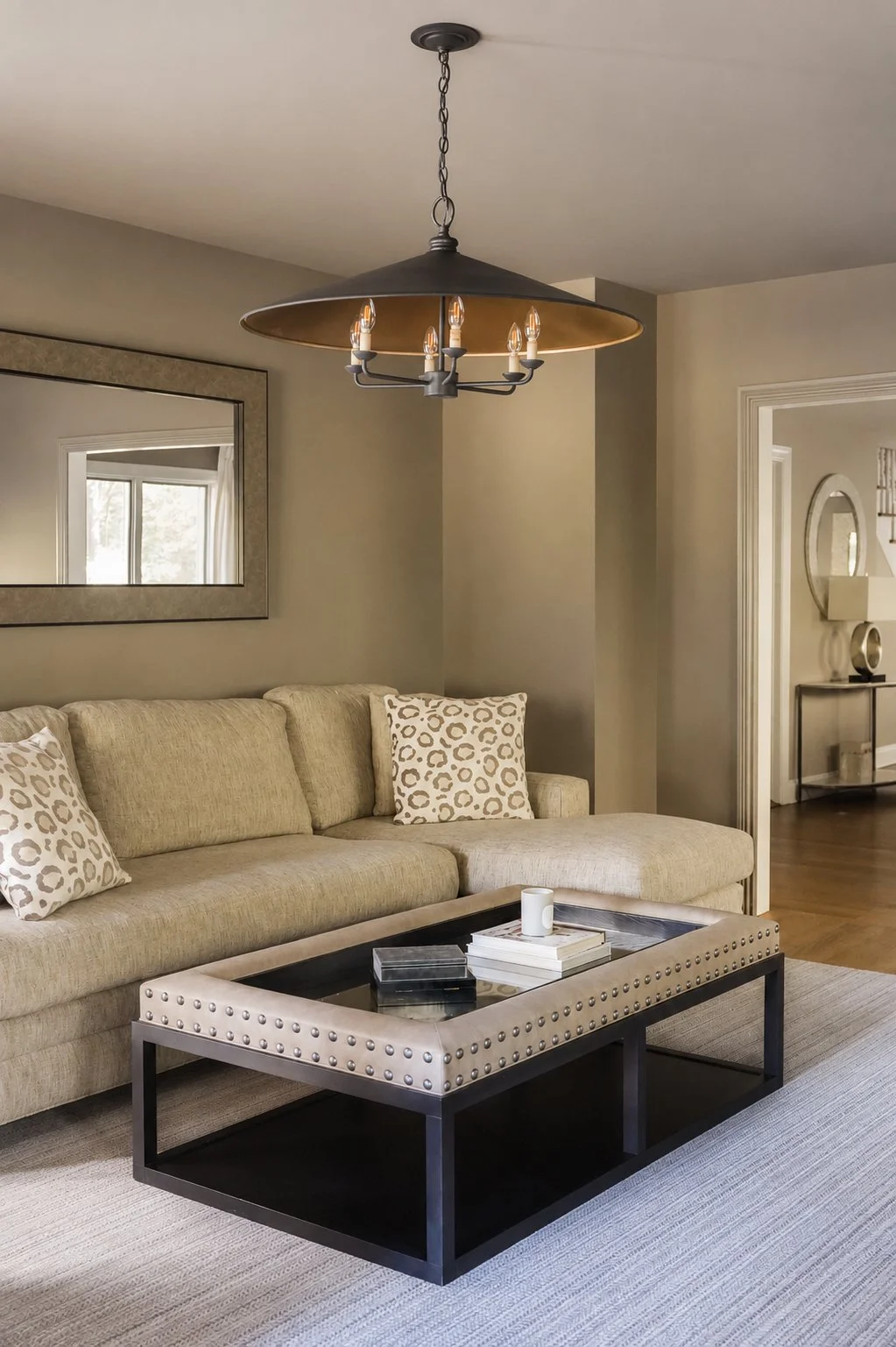 Living room with beige sofa, patterned pillows, black coffee table with books and a mug, a large mirror on the wall, beige walls, and a ceiling light fixture with six bulbs hanging above.
