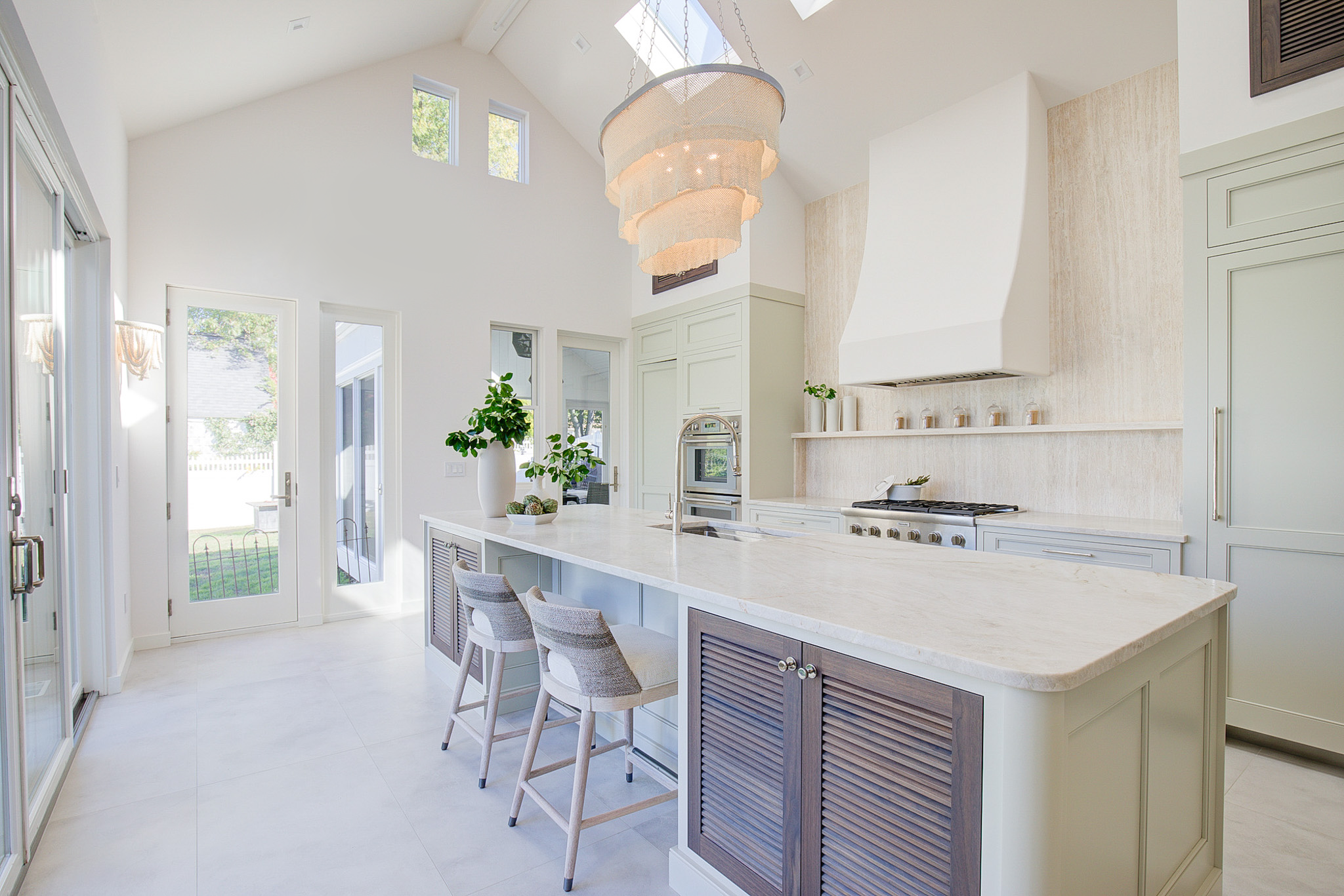 Bright white kitchen with large island, modern appliances, and a chandelier, with multiple windows and glass door letting in natural light.