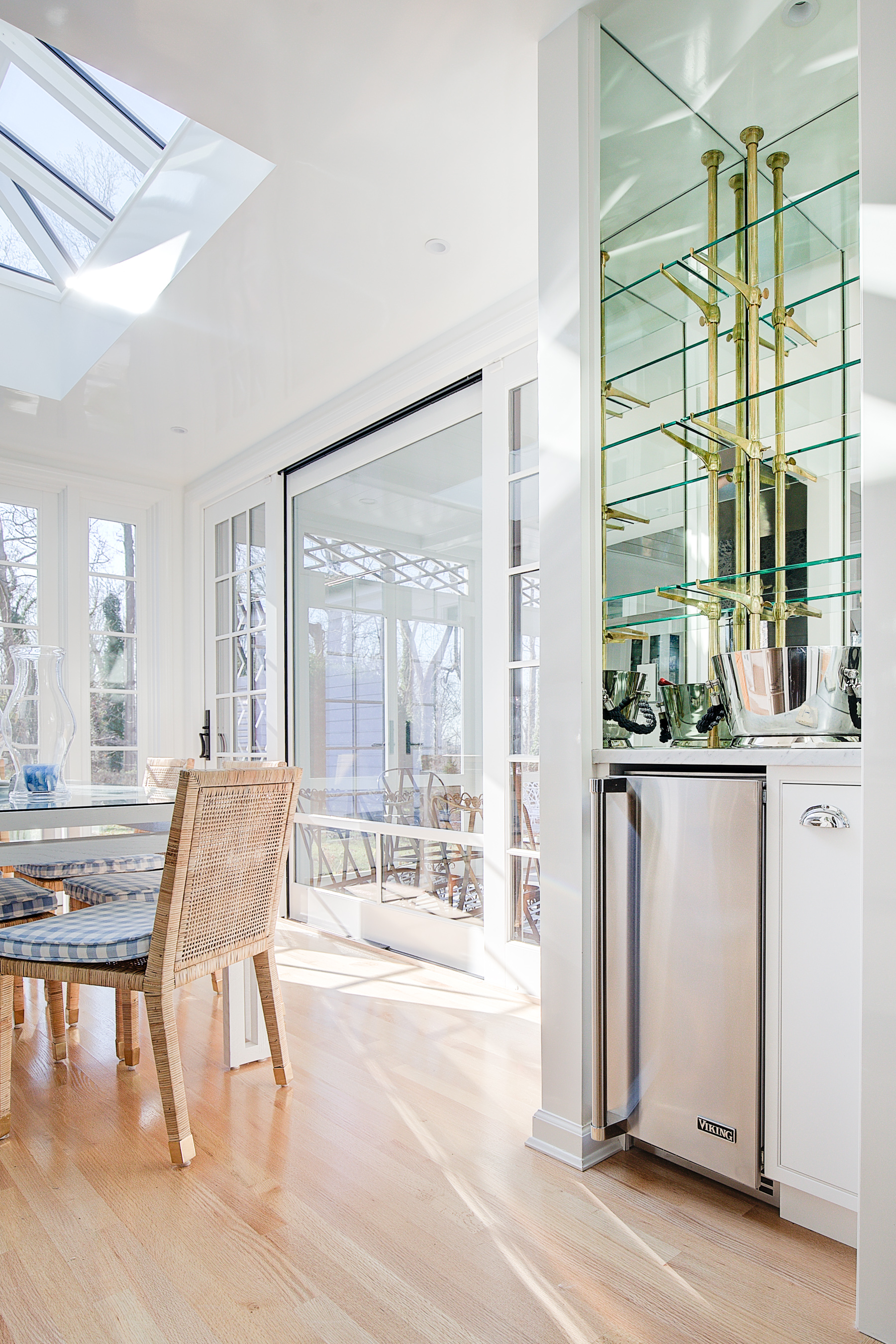 Bright sunlit dining area with glass door and windows, wooden floor, wicker and cushioned chairs, and a mirrored display with gold accents.