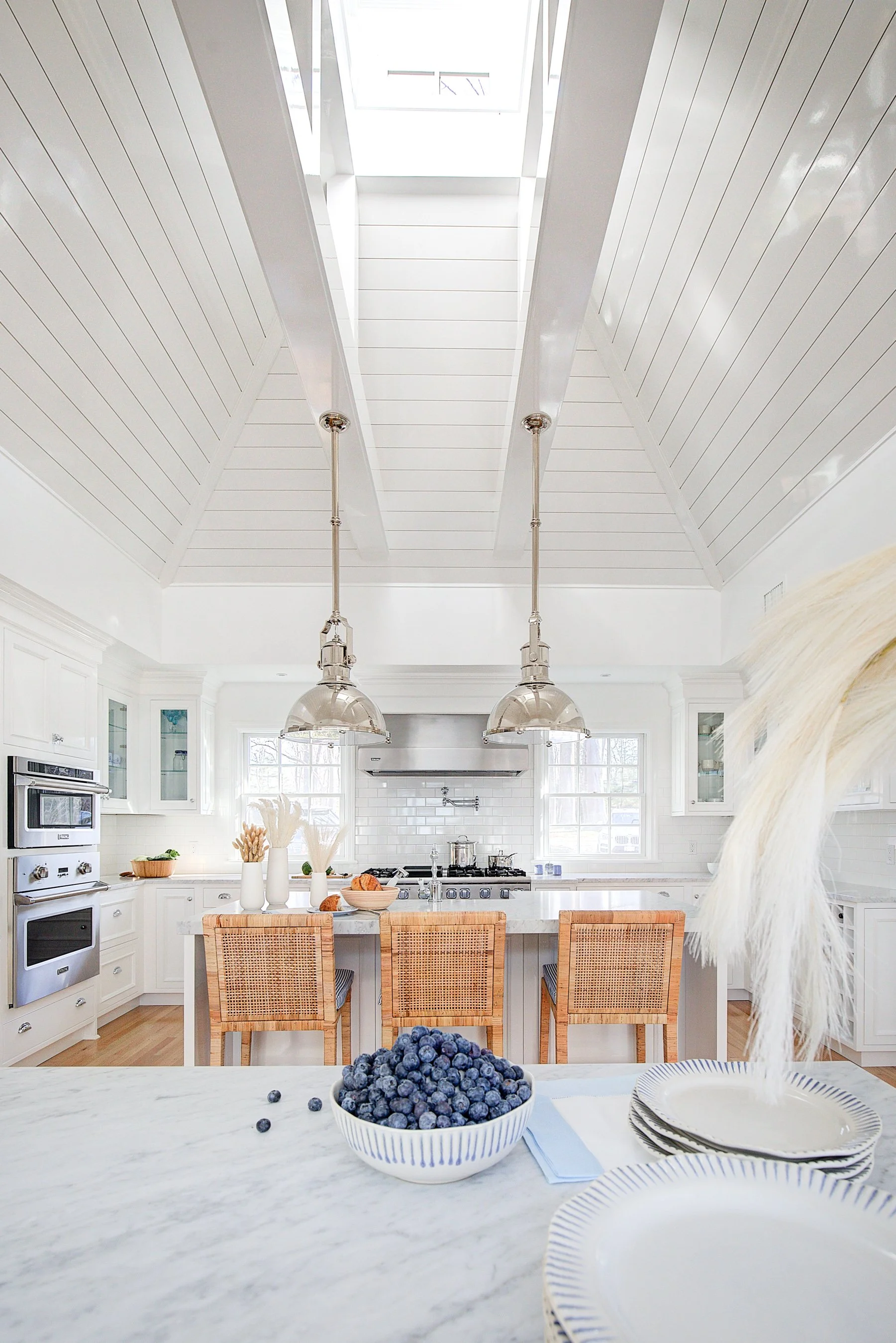 A bright, white kitchen with a skylight ceiling, white cabinets, and a marble island. On the island, there are bowls of blueberries and plates with striped rims. In the background, there is a dining area with wicker chairs, and the kitchen has hanging pendant lights above the island.