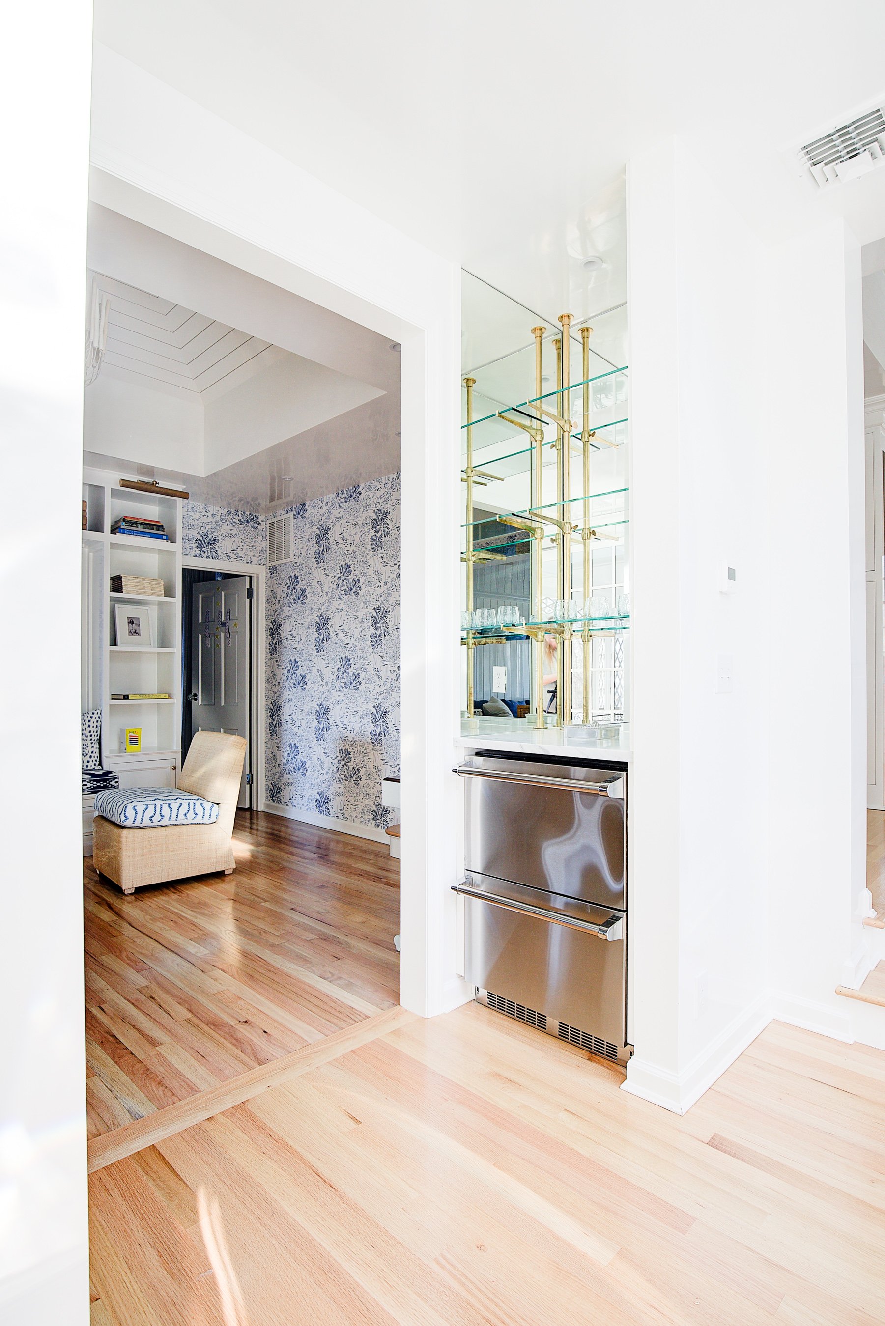 Open doorway leading into living space with hardwood flooring, a blue and white wallpapered wall, a white bookshelf with books and decorative items, a beige armchair, and a closet door.
