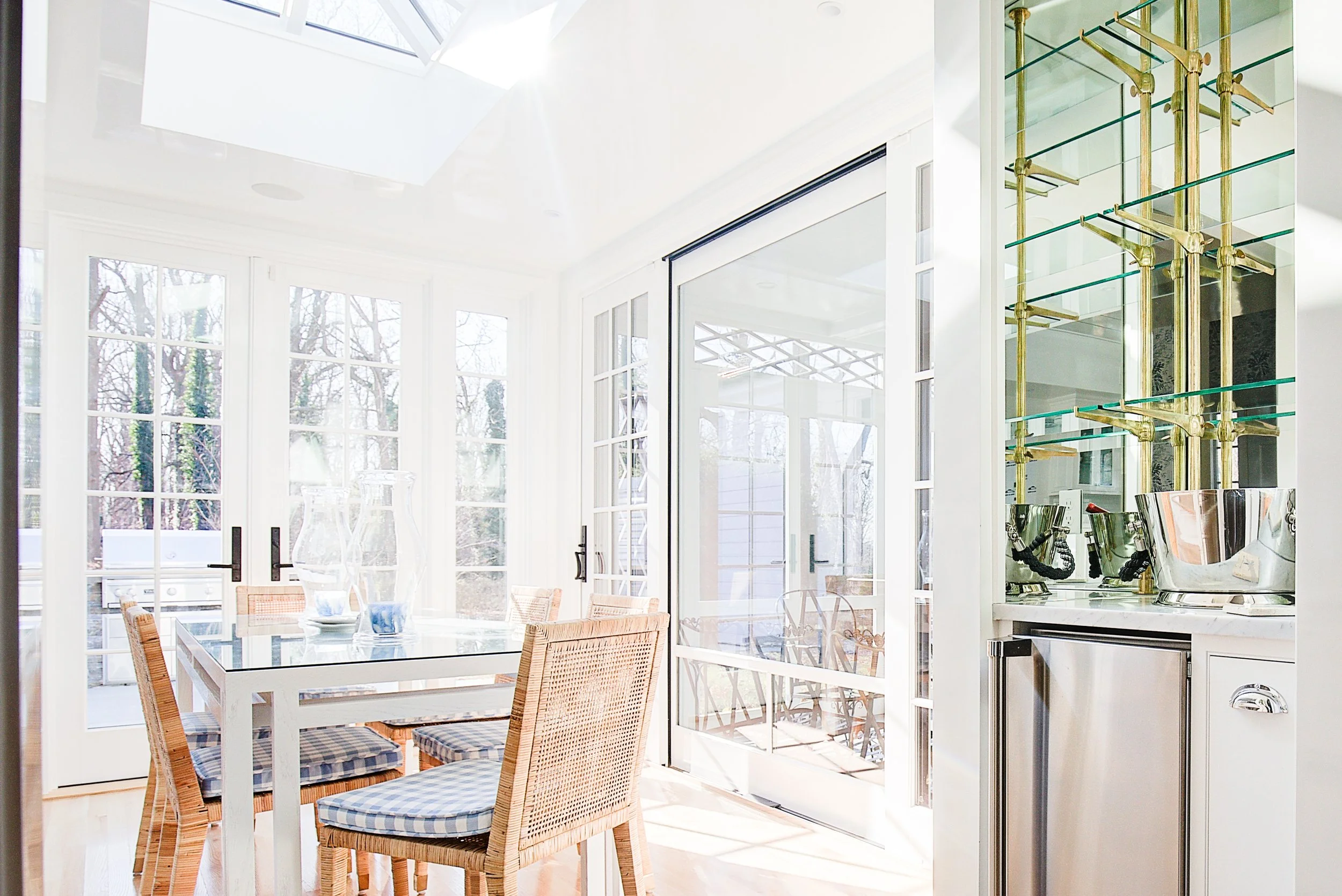 Bright dining area with glass sliding doors, wooden dining table, and rattan chairs with checked cushions, adjacent to a kitchen with glass shelves and brass fixtures.