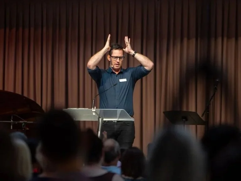 An adult male standing on a stage in front of an audience, with his hands making a crown shape above his head