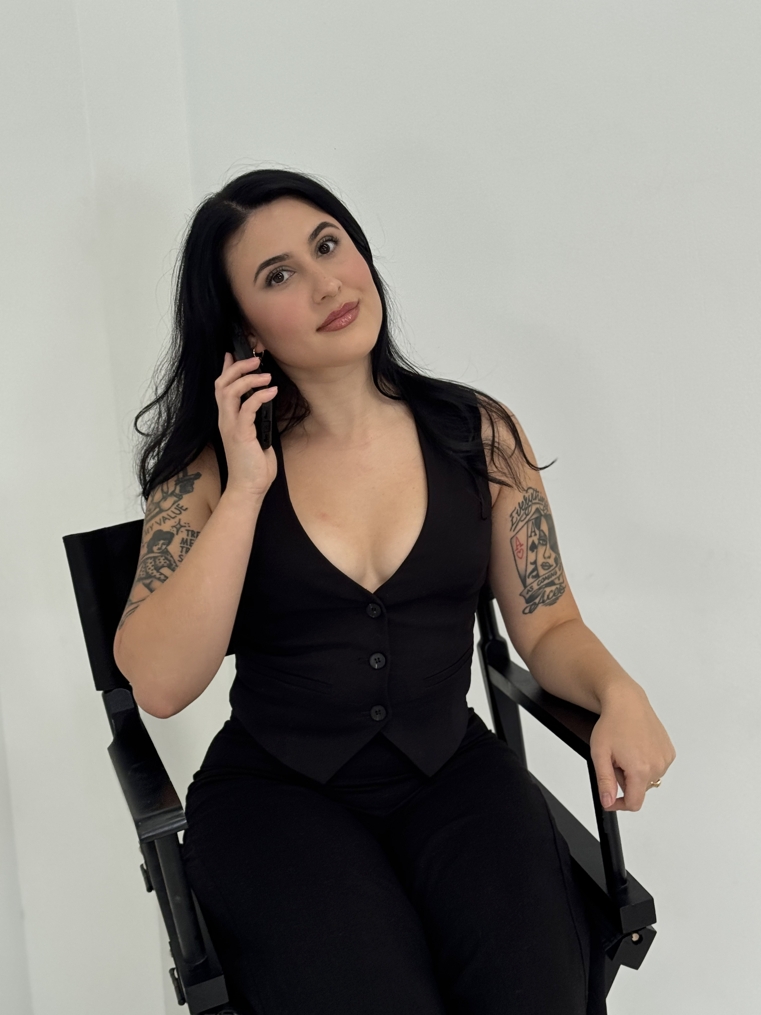 A woman with black hair and tattoos on her arms sitting on a black chair, talking on a phone, wearing a black sleeveless top and black pants, against a plain white wall.