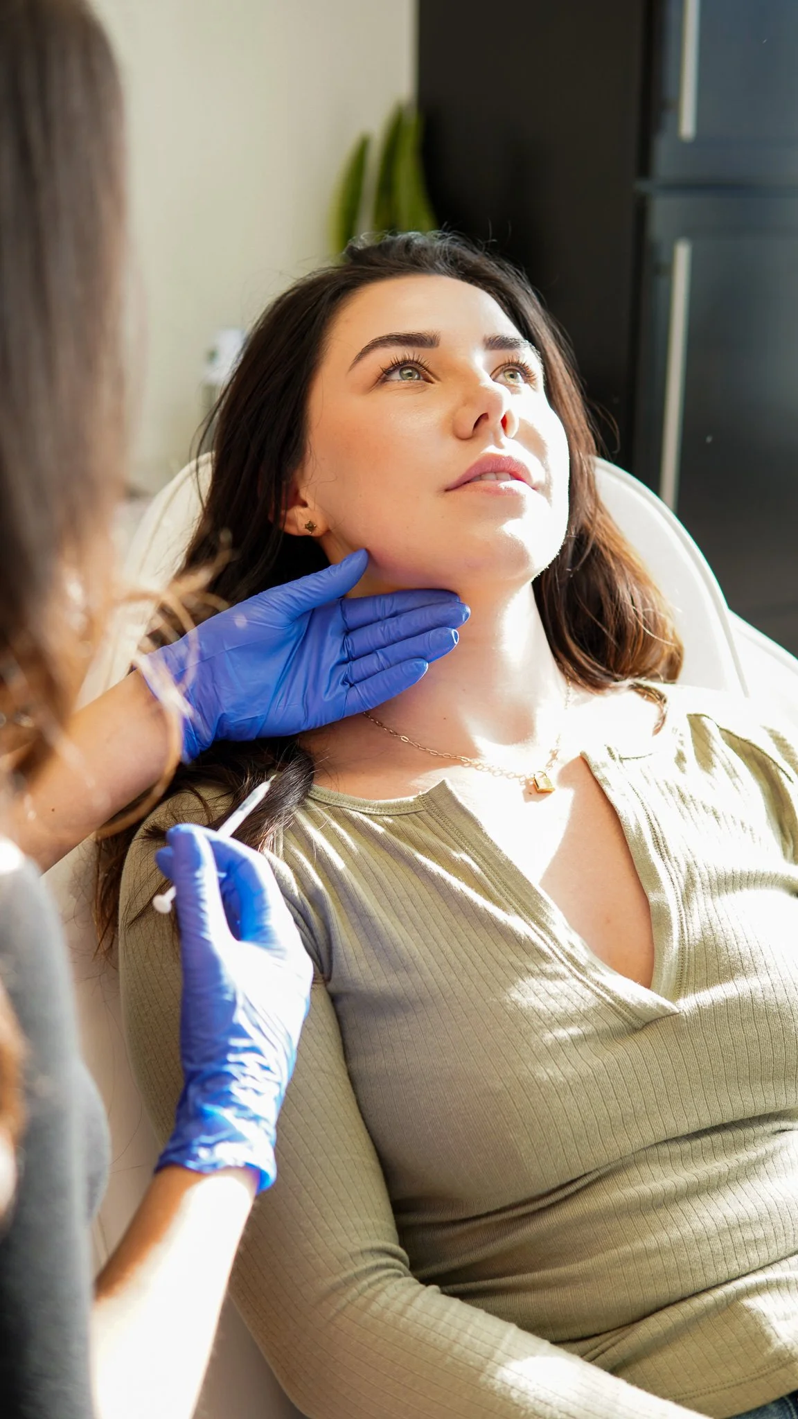 A woman with dark hair and light skin lying back while receiving a medical shot in her upper arm, with a healthcare professional wearing blue gloves holding the syringe and supporting her neck.