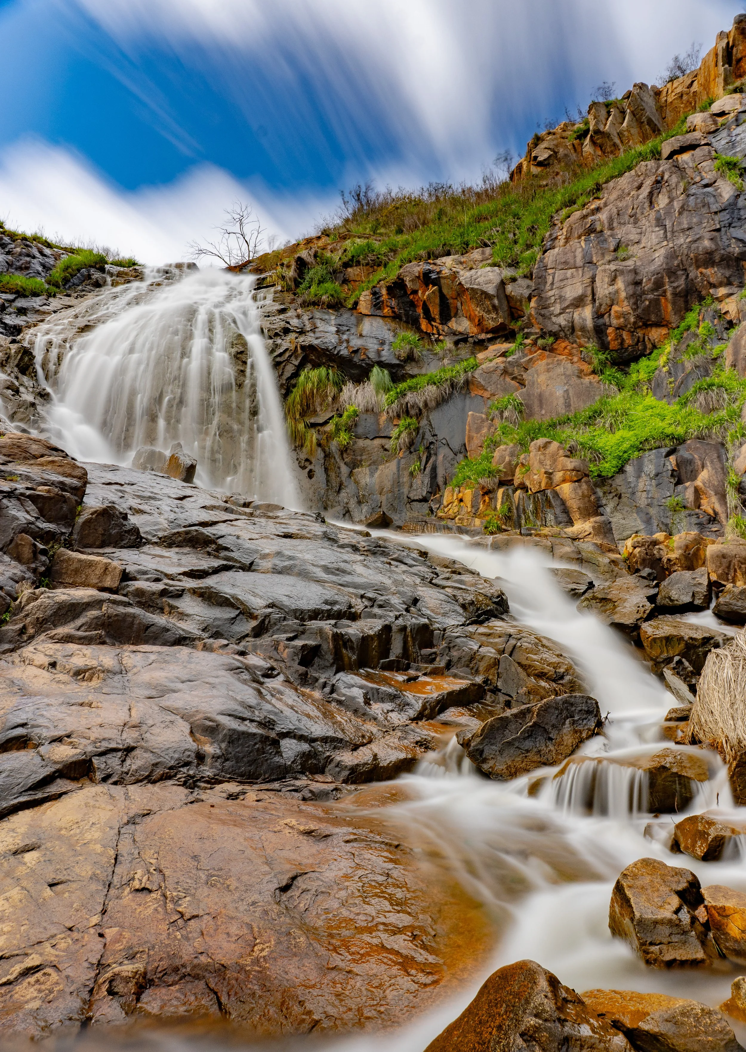 A waterfall cascading down rocky cliffs surrounded by green grass and moss under a partly cloudy sky.