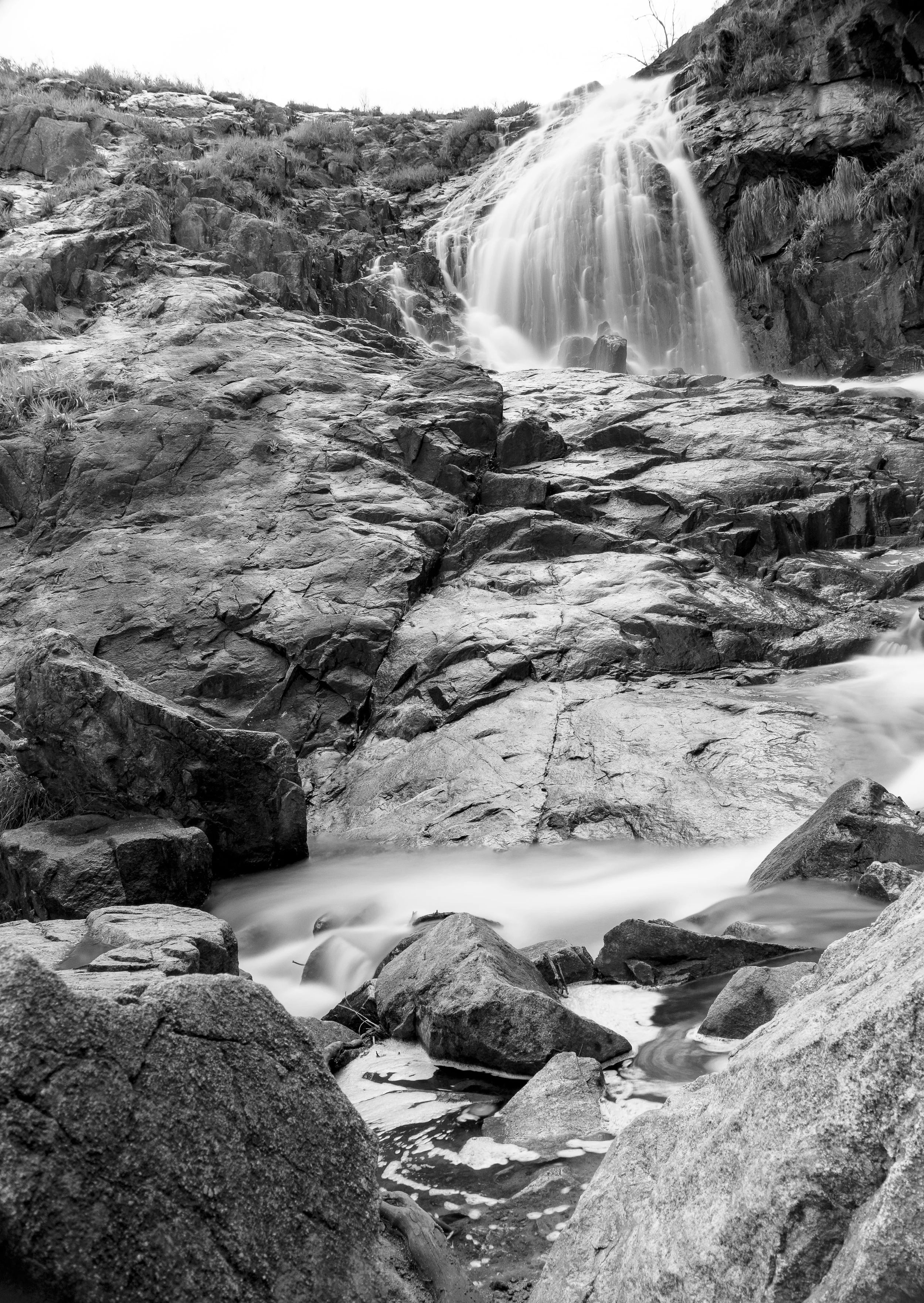 Black and white photo of a waterfall cascading over rocks, flowing into a stream surrounded by large rocks and boulders.