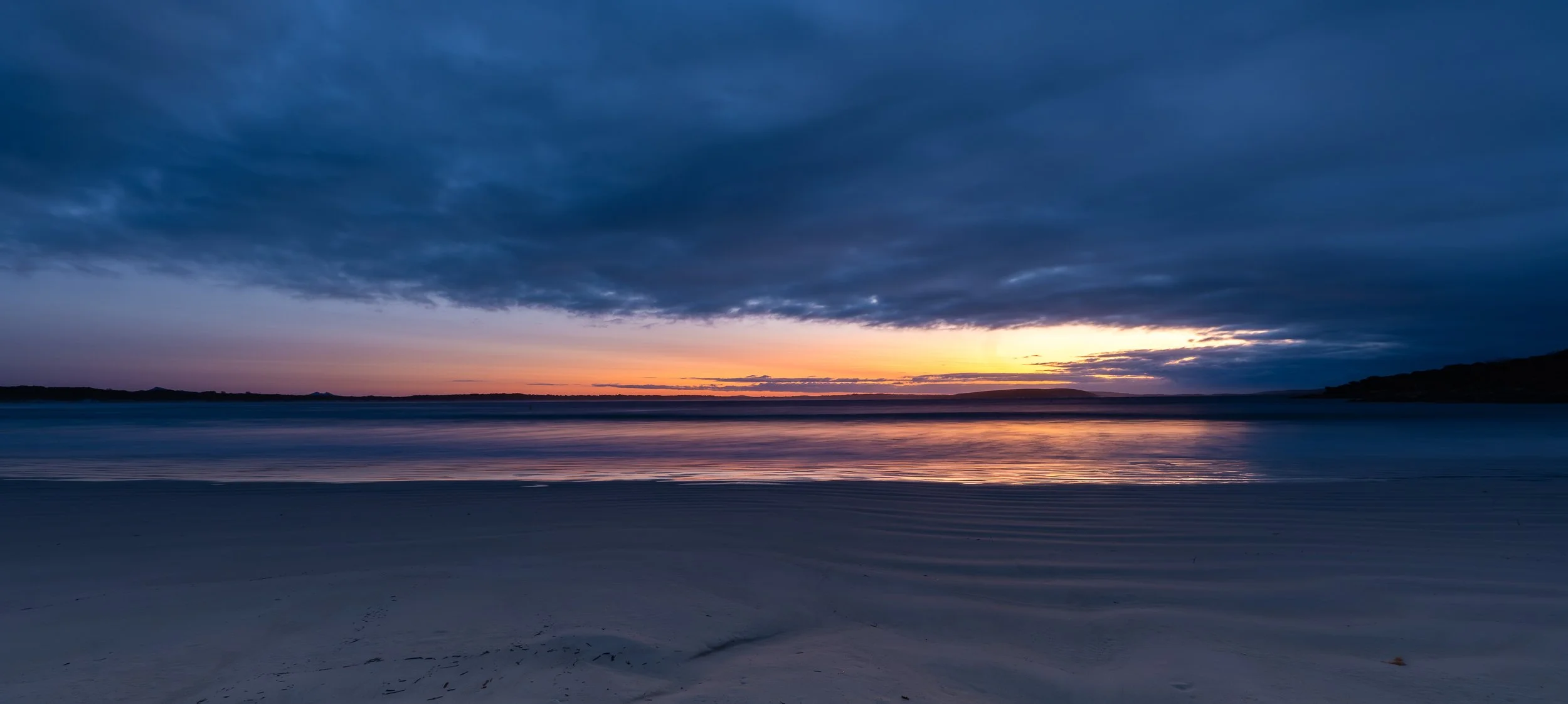 A serene beach at sunset with a calm body of water, a sandy shoreline, and a dark cloudy sky with a hint of orange and purple near the horizon.