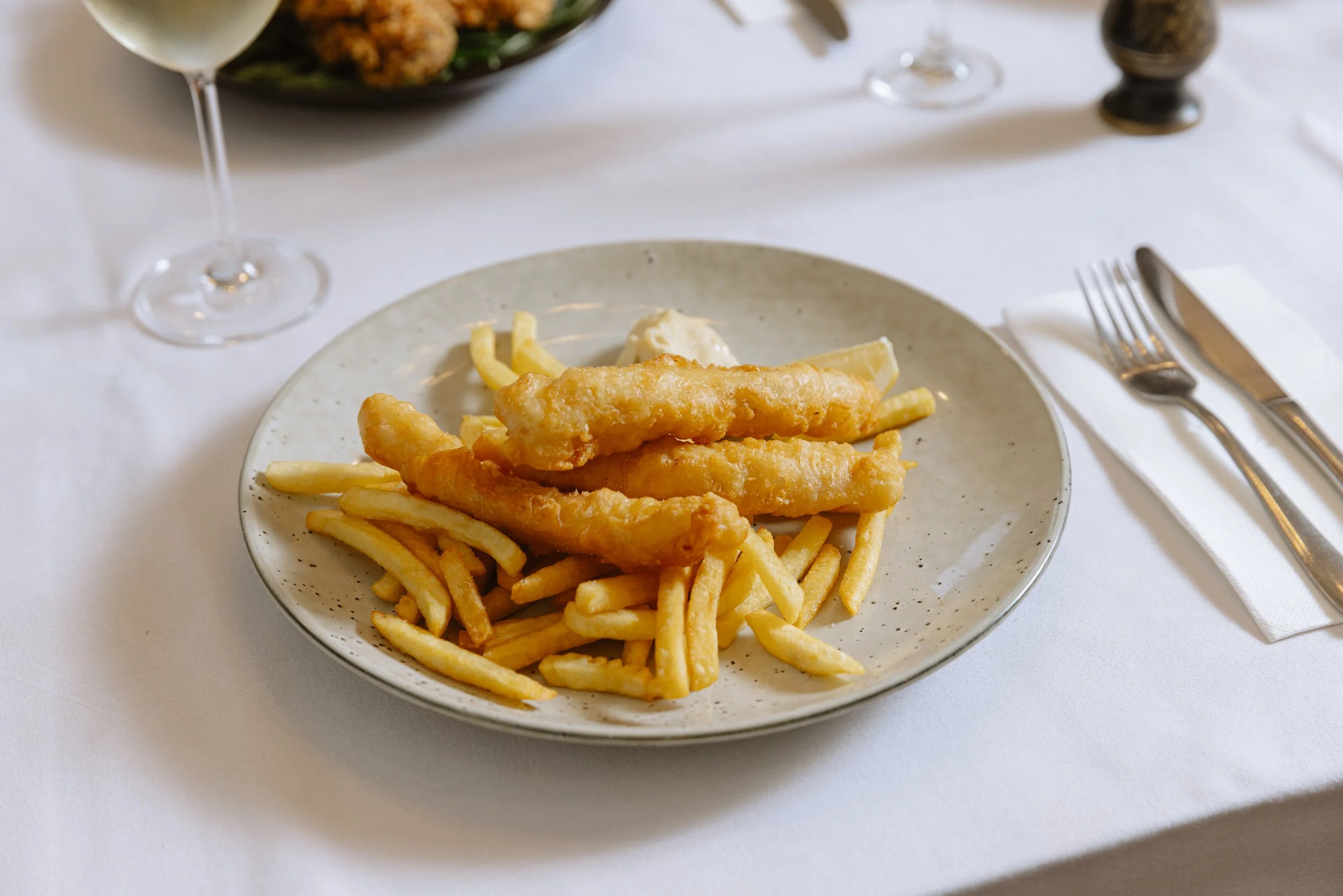 Plate of fried fish fillets with French fries on a white tablecloth, with a glass of white wine and a salad in the background at The Mint pub melbourne.
