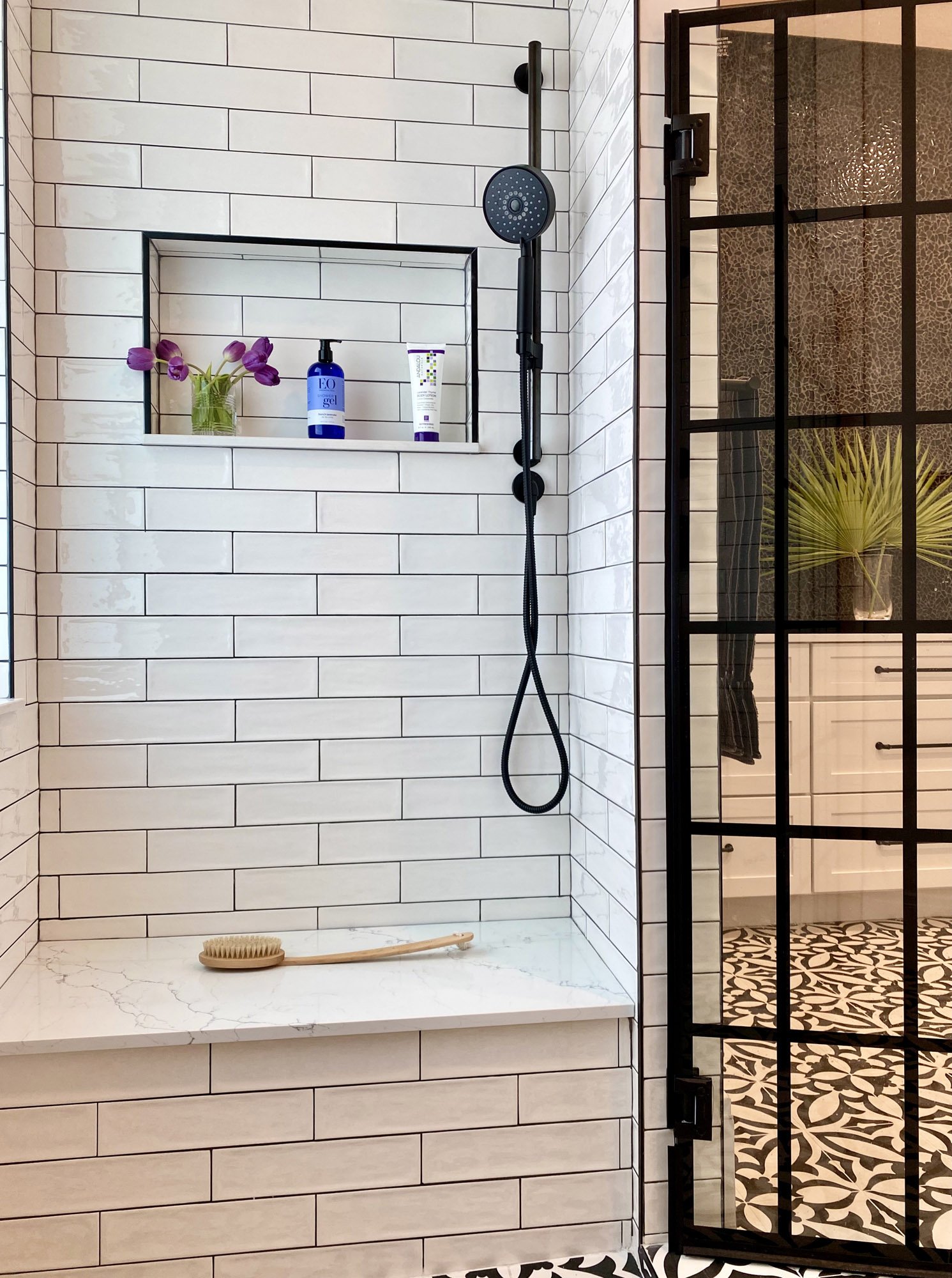 Wellington, Florida master bedroom shower room with white subway tile, built in bench, black shower head, black glass shower door. View into bathroom shows tile and custom vanity.