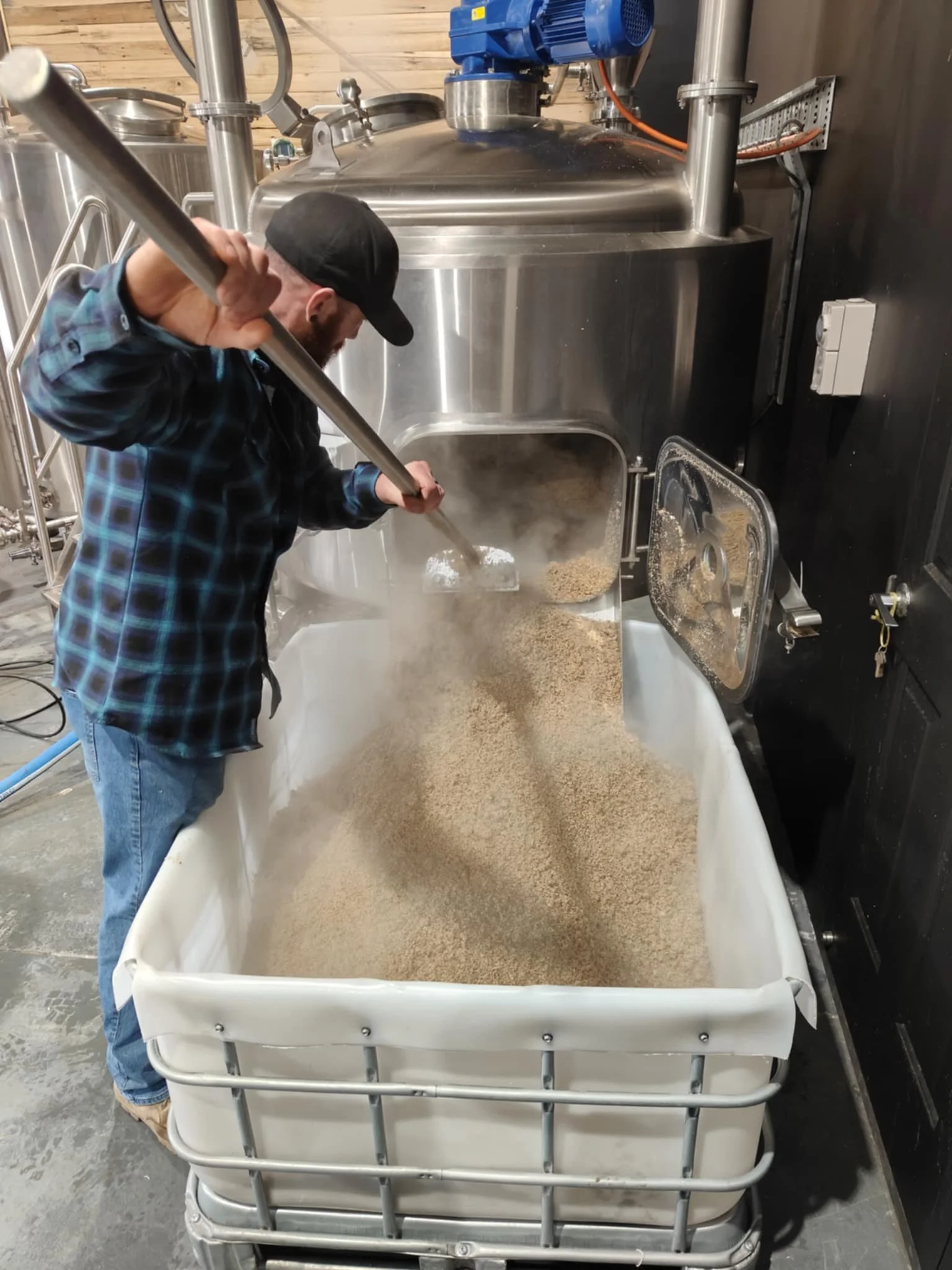 A man in a plaid shirt and black cap stirring grains in a large stainless steel brewing tank in a brewery.