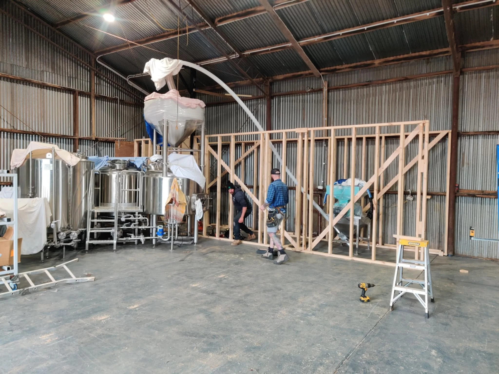 Construction workers inside a large industrial space building a wooden wall frame with stainless steel brewing equipment visible on the left.