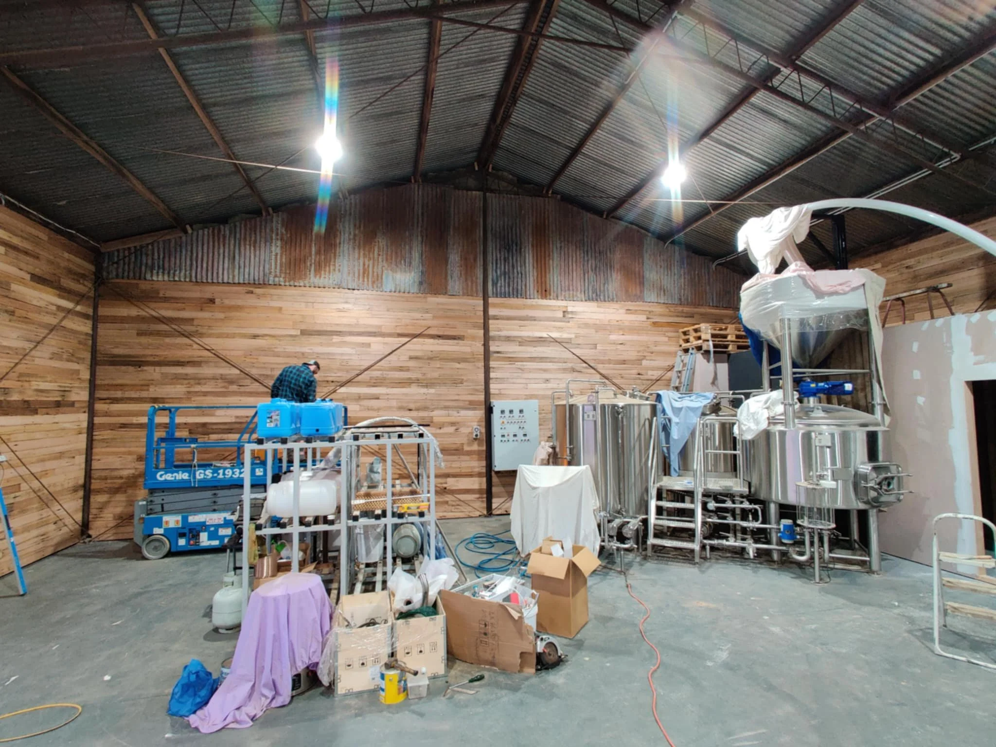 Interior of a brewery or processing facility with metal tanks, equipment, and scattered supplies; a person is working on a blue lift platform against a wooden wall, with tools and boxes on the floor.