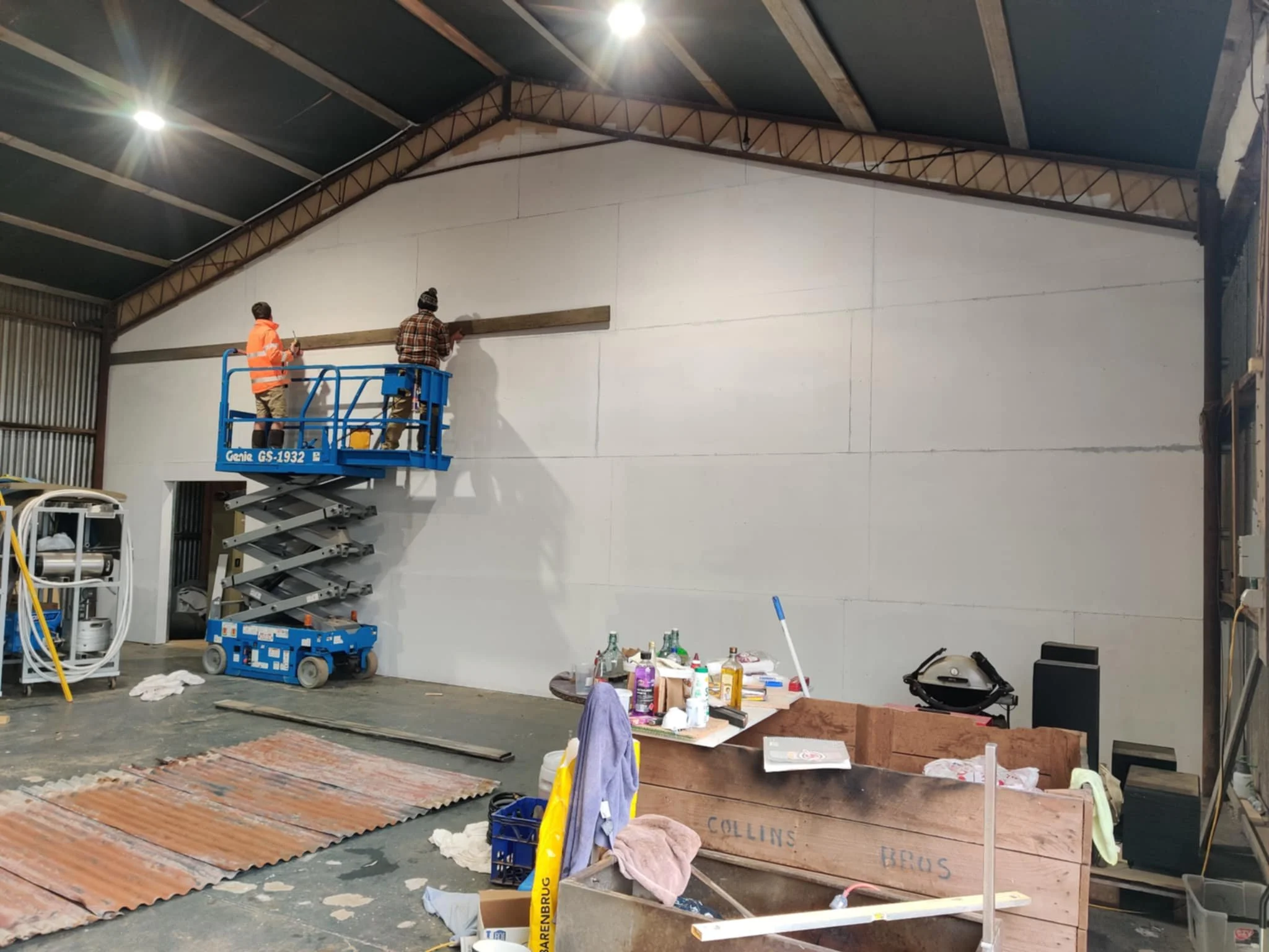 Two workers are on a blue scissor lift working on the high white wall inside a warehouse or workshop. One worker is wearing an orange safety vest, and the other is wearing a beanie and plaid shirt. The workspace has various tools, supplies, and build