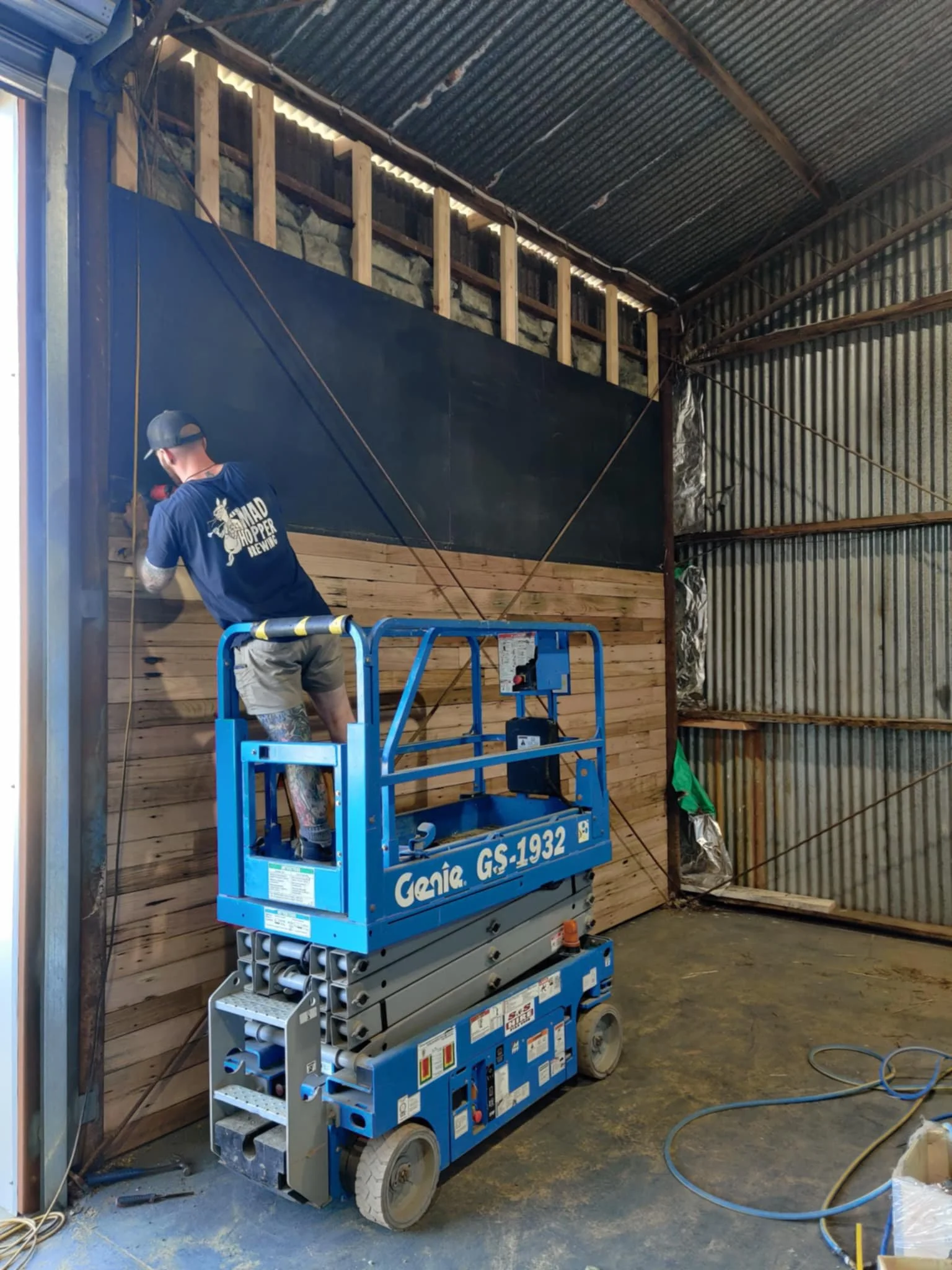 A worker in a navy blue t-shirt with a bee graphic and the words 'HOPPER HEW' is using a power tool to attach wooden panels to a wall inside a metal warehouse. The worker is standing on a blue Genie GS-1932 scissor lift, which is parked on a concrete