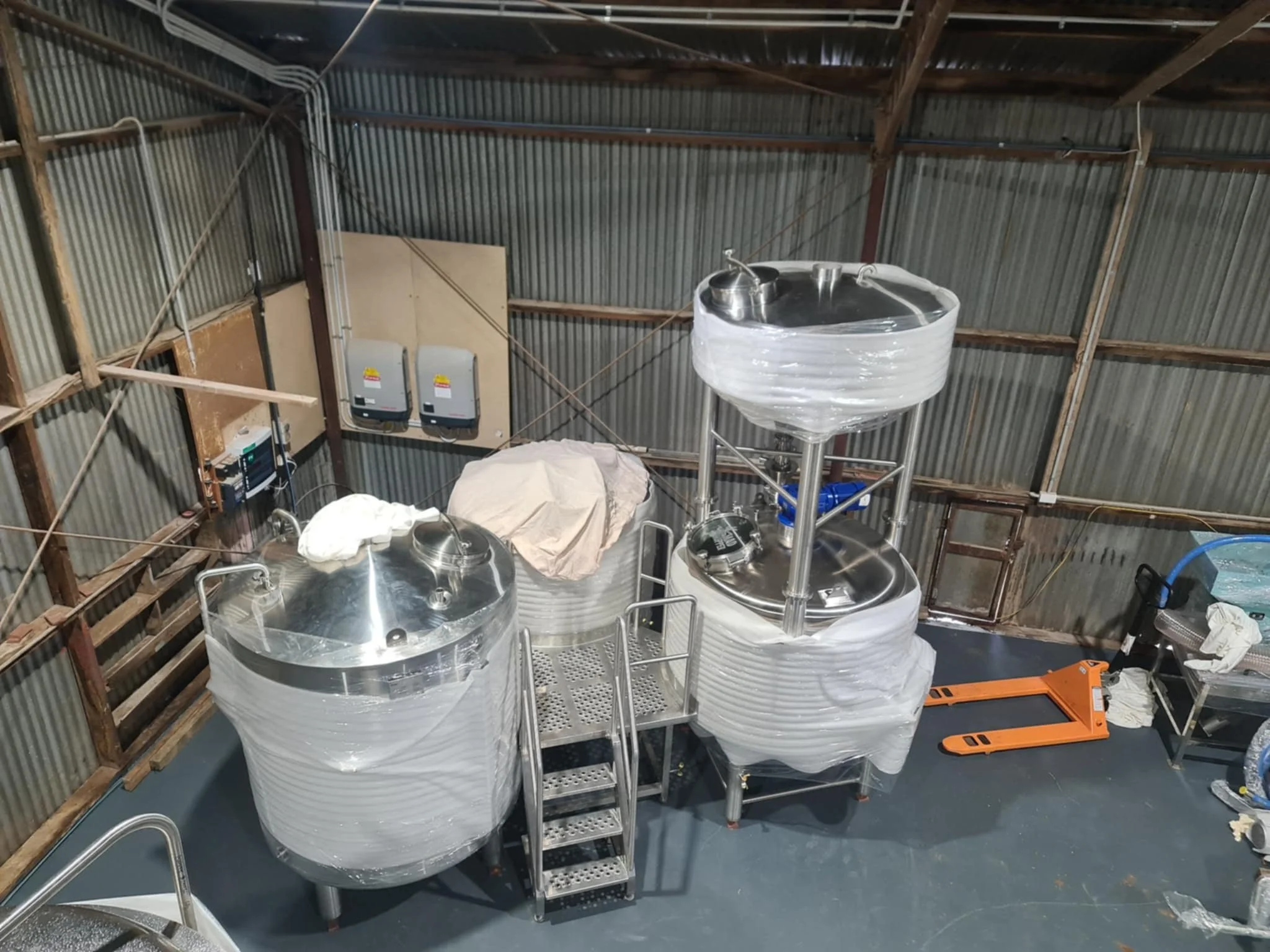 Industrial brewing equipment with large stainless steel tanks wrapped in plastic, set inside a metal warehouse with exposed corrugated metal walls and a wooden roof structure.
