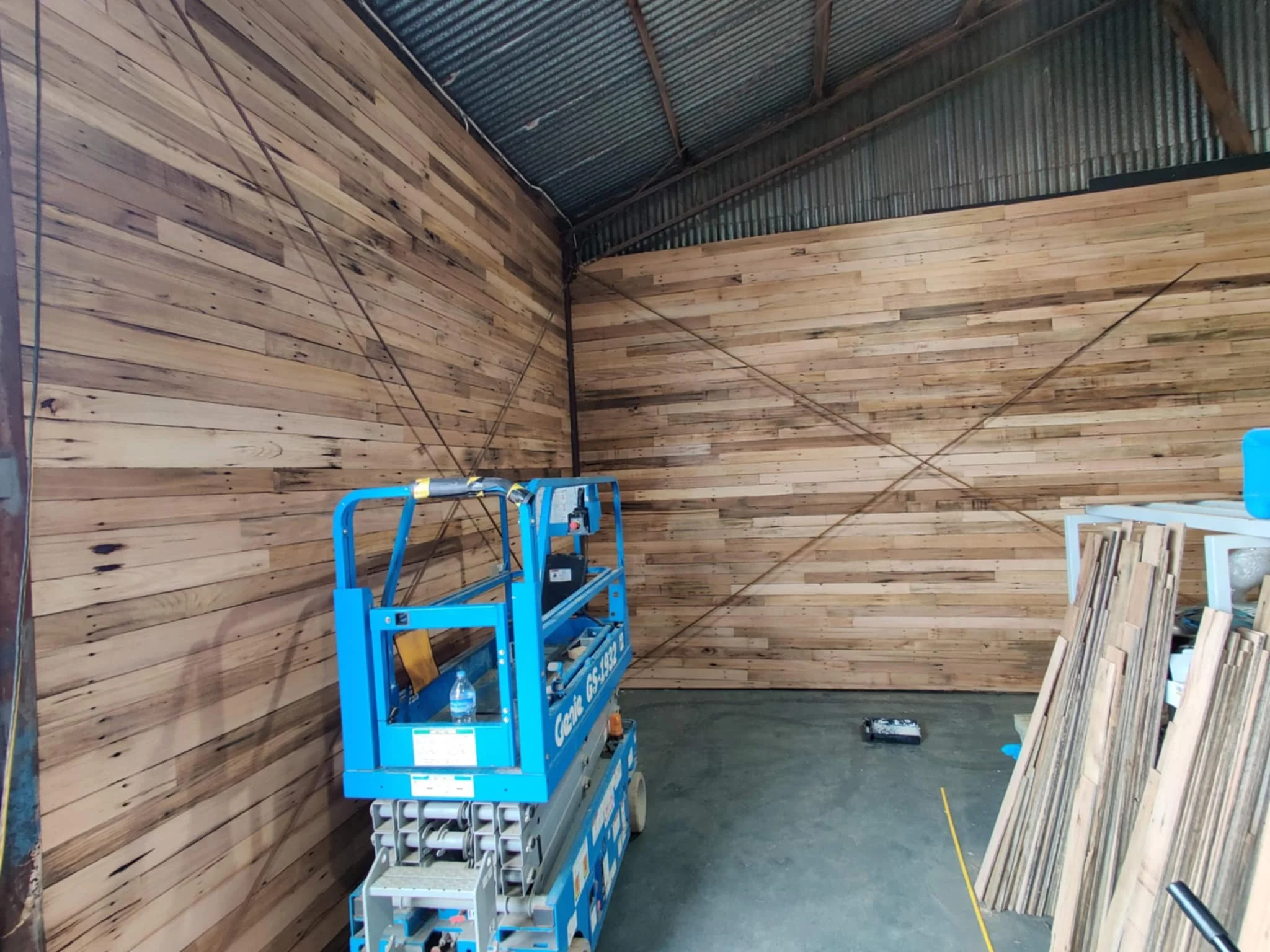 Interior of a woodworking workshop with wooden paneled walls and a steel roof, featuring a blue scissor lift, a water bottle on it, and stacks of wood on the right side.
