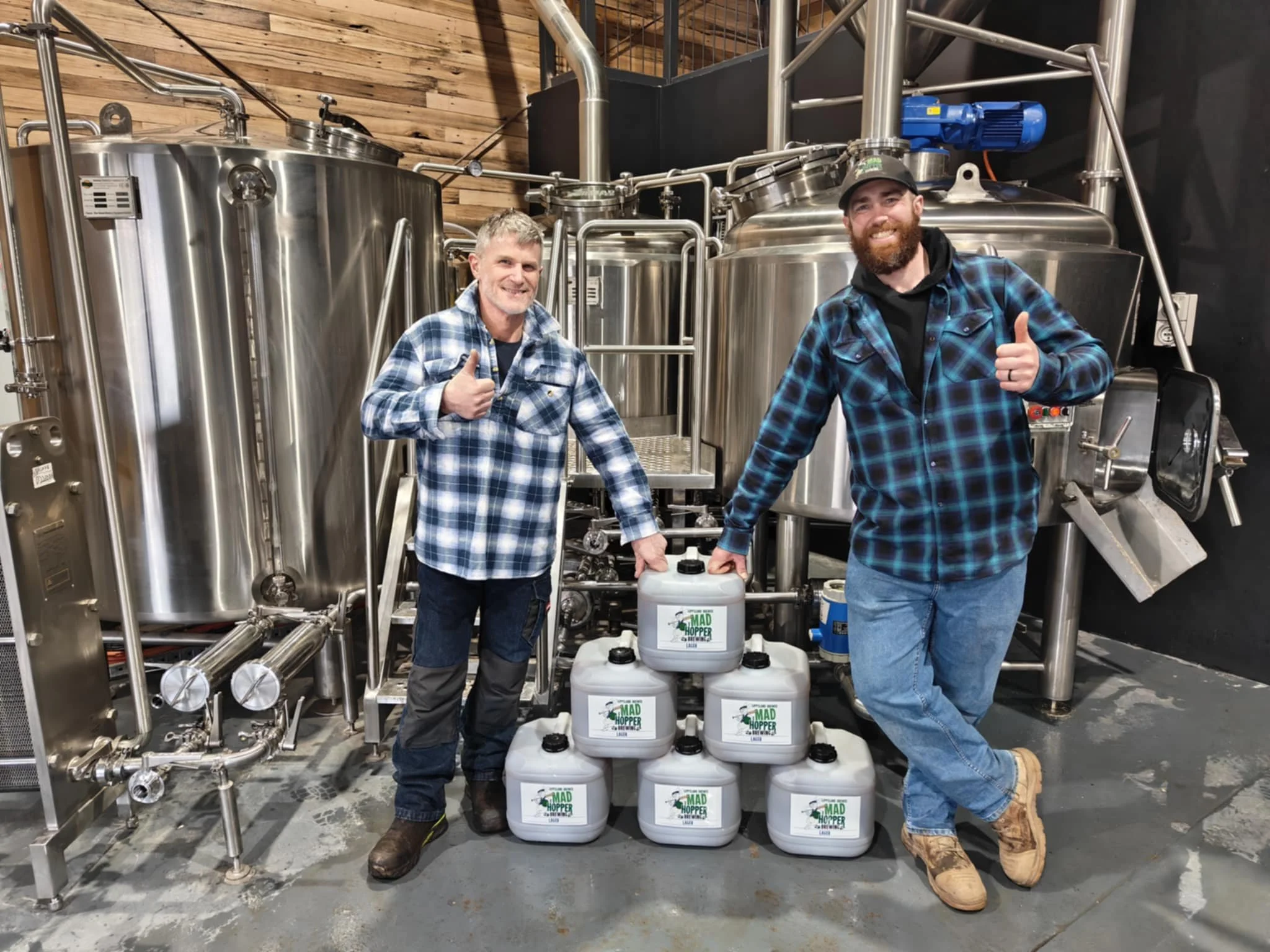 Two men in plaid shirts and jeans standing in front of large stainless steel brewing equipment, smiling and giving thumbs up. There are six white plastic containers labeled 'MAD HOPPER' stacked on the floor between them.