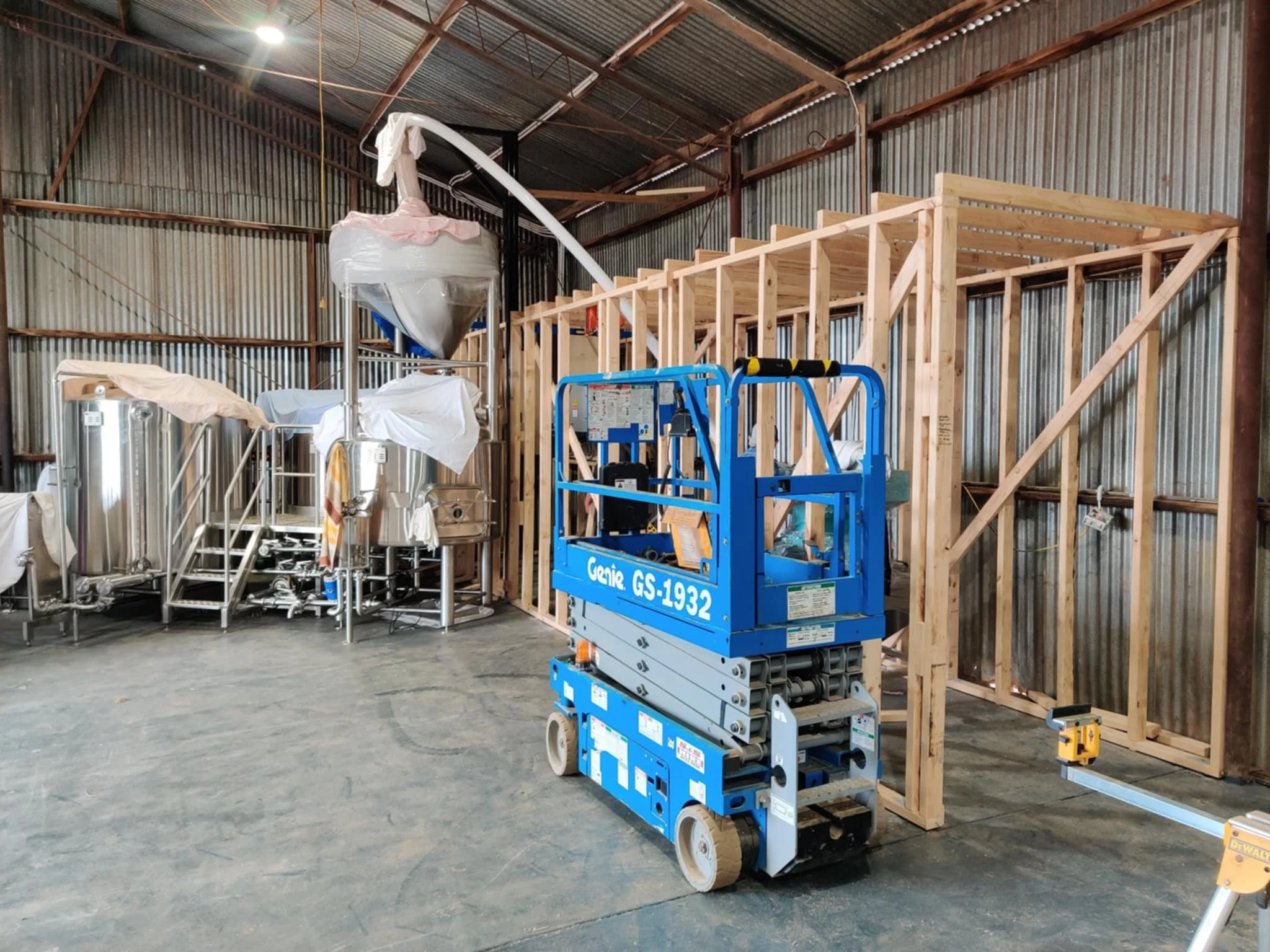 Construction inside a metal building with wooden framing, construction lift, and stainless steel equipment covered with plastic.
