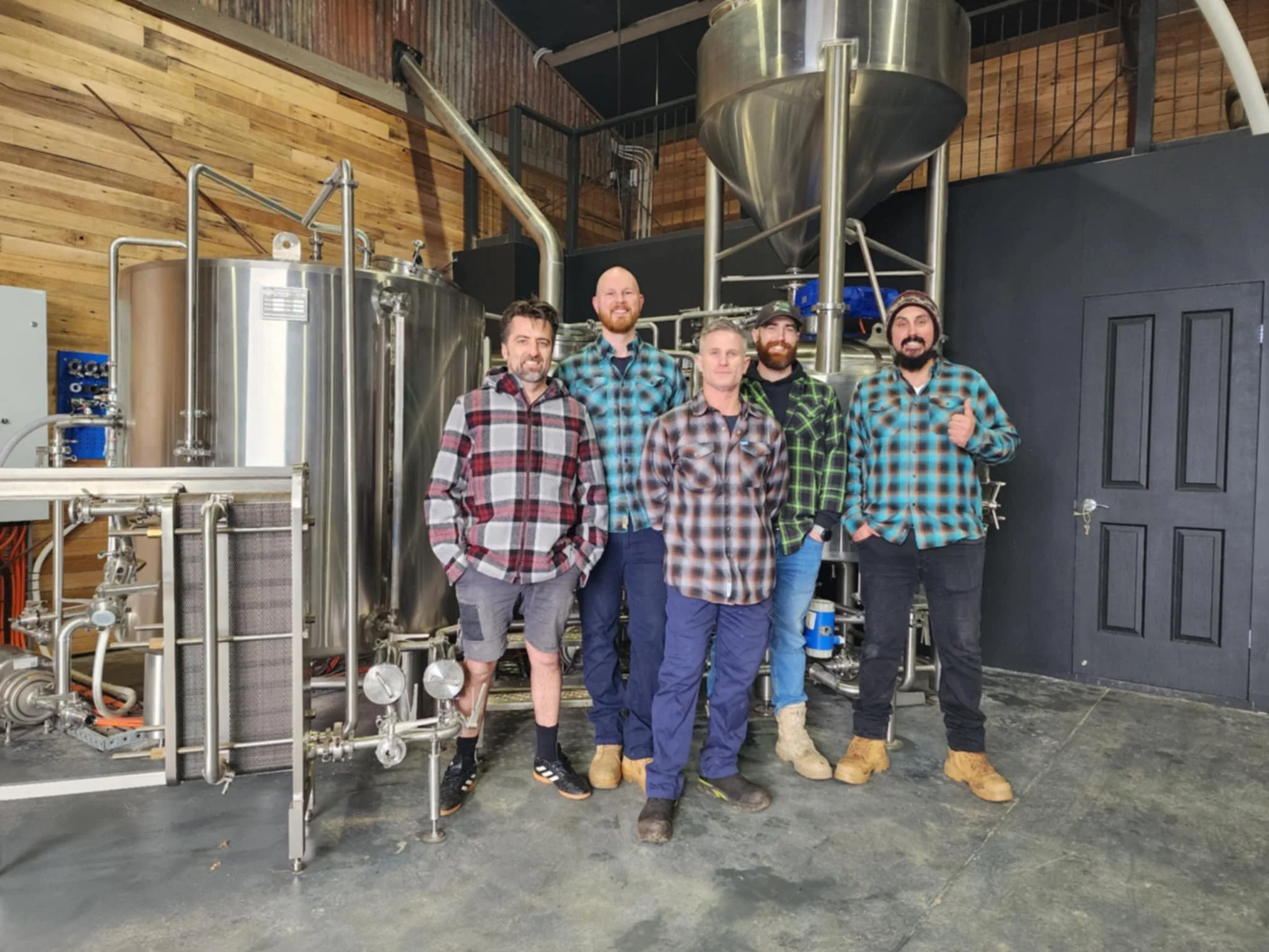 Five men standing in front of stainless steel brewing equipment in a brewery, wearing plaid shirts and jeans or shorts, with a wooden wall and black door in the background.