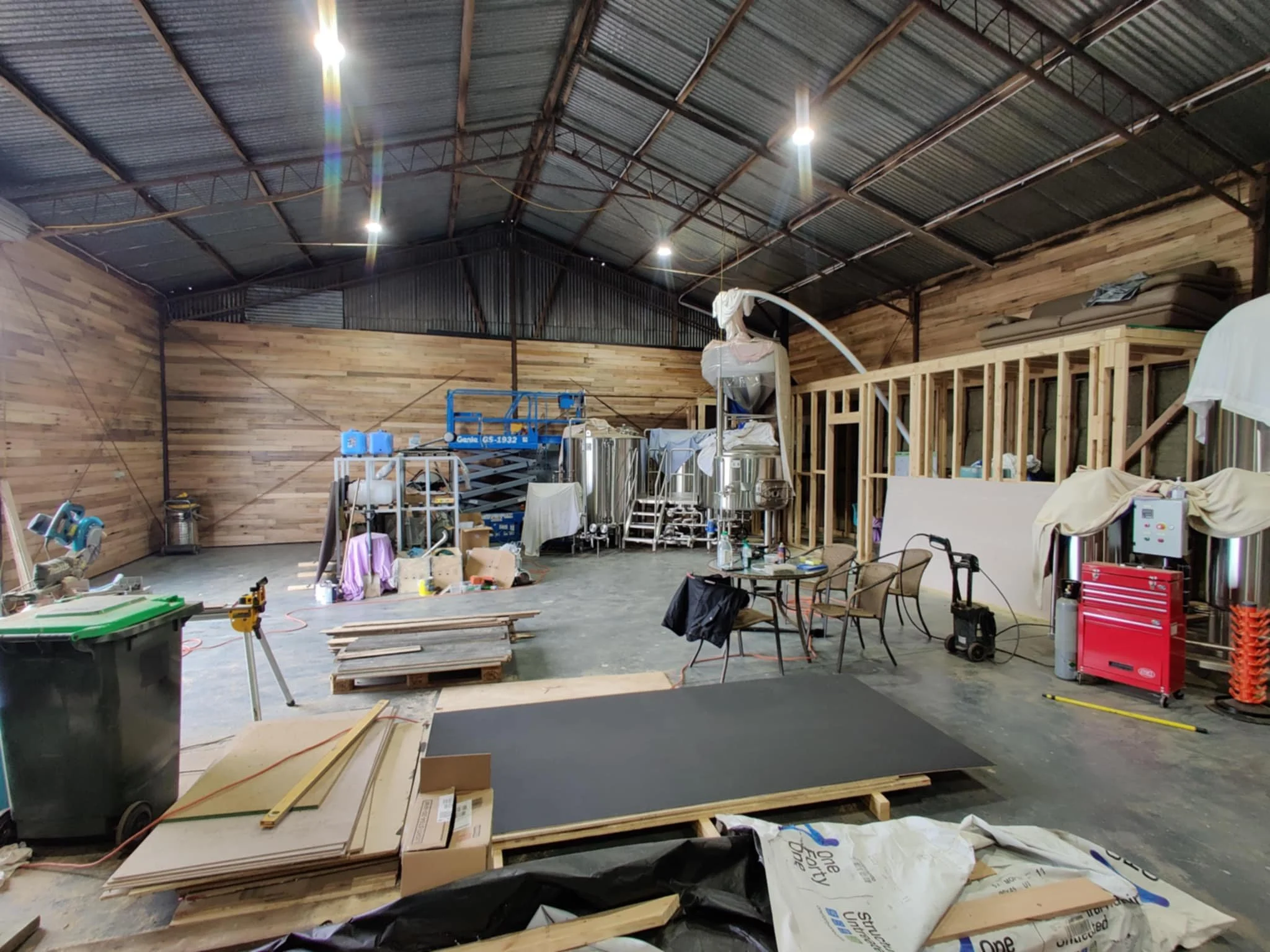 Interior of a workshop or brewery under construction with wood panel walls, metal roof, and various equipment, tools, and furniture scattered around.