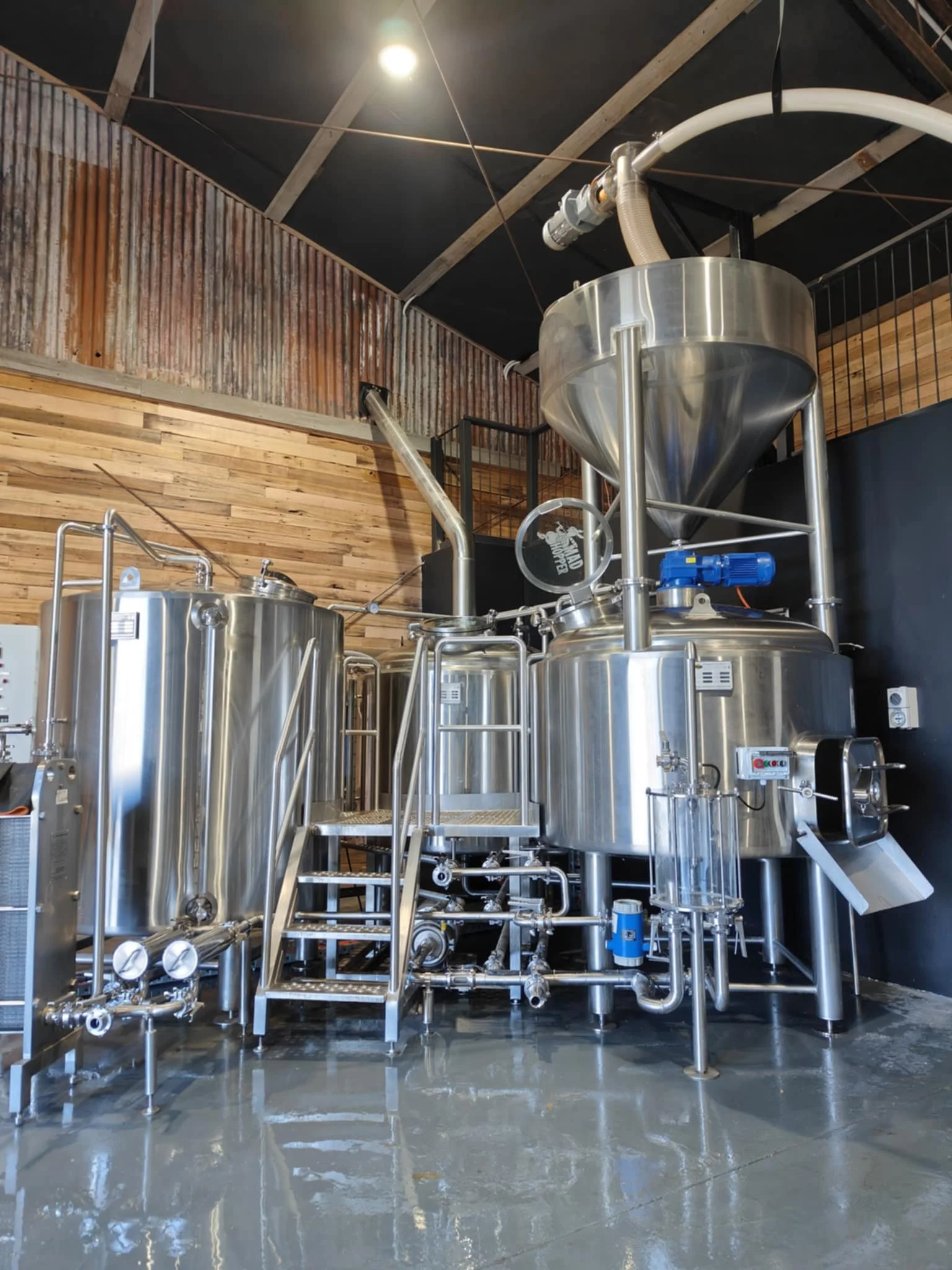 Industrial stainless steel brewing equipment in a brewery with wooden wall panels and a black ceiling.