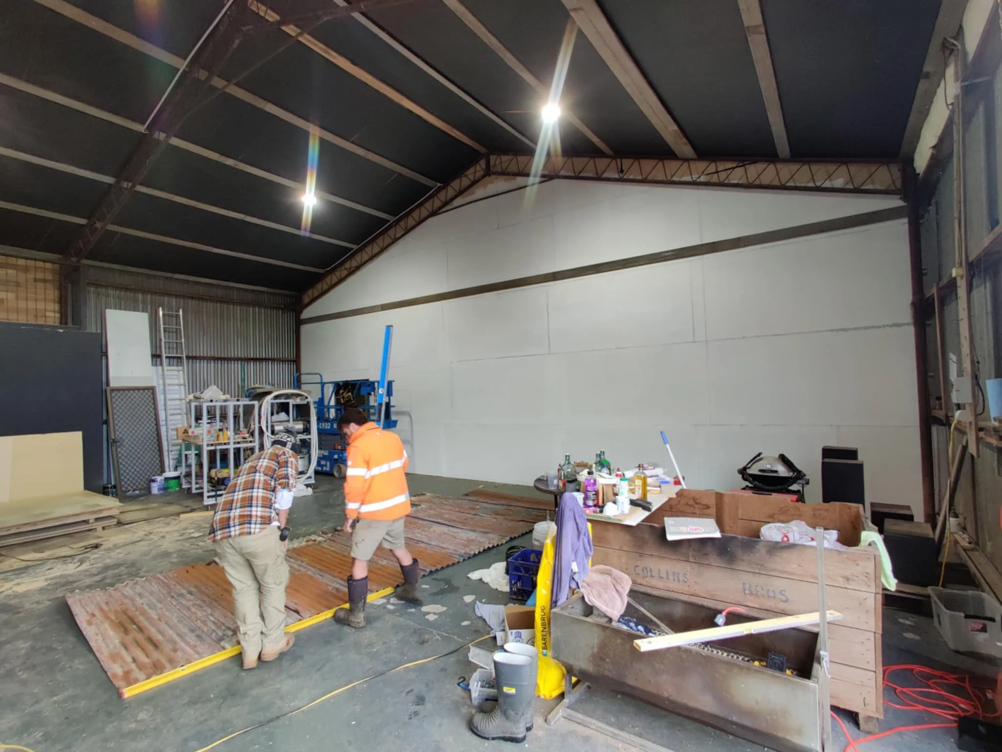 Two workers in an indoor construction or renovation space, measuring a corrugated metal sheet on the floor. The area contains tools, supplies, and storage shelves, with a partially finished wall.