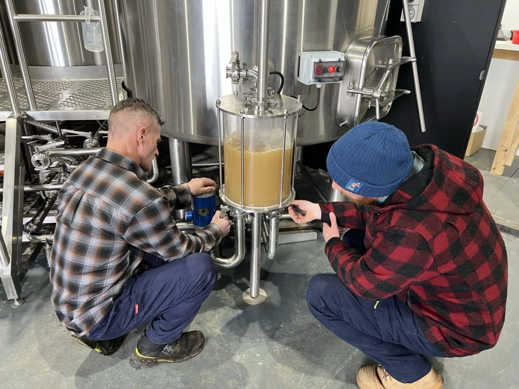 Two people working on a stainless steel brewing tank, one kneeling and the other crouching, with brewing equipment around them.
