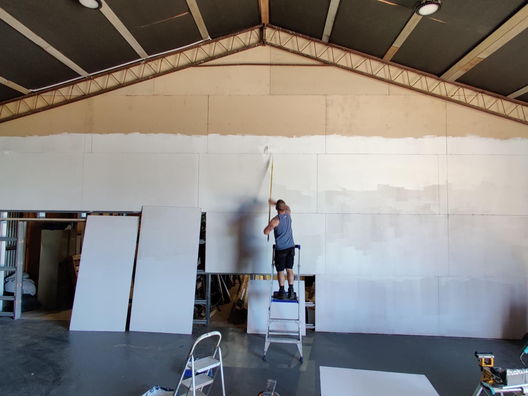 A person standing on a step ladder using a tape measure to measure a large wall under construction or renovation inside a building with a high ceiling and exposed beams.
