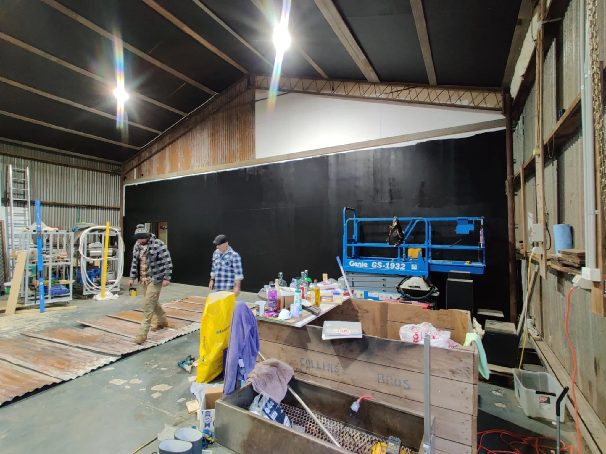 Inside a warehouse with two men working; one in a checkered jacket and another in a blue plaid shirt, surrounded by tools, equipment, and construction materials.