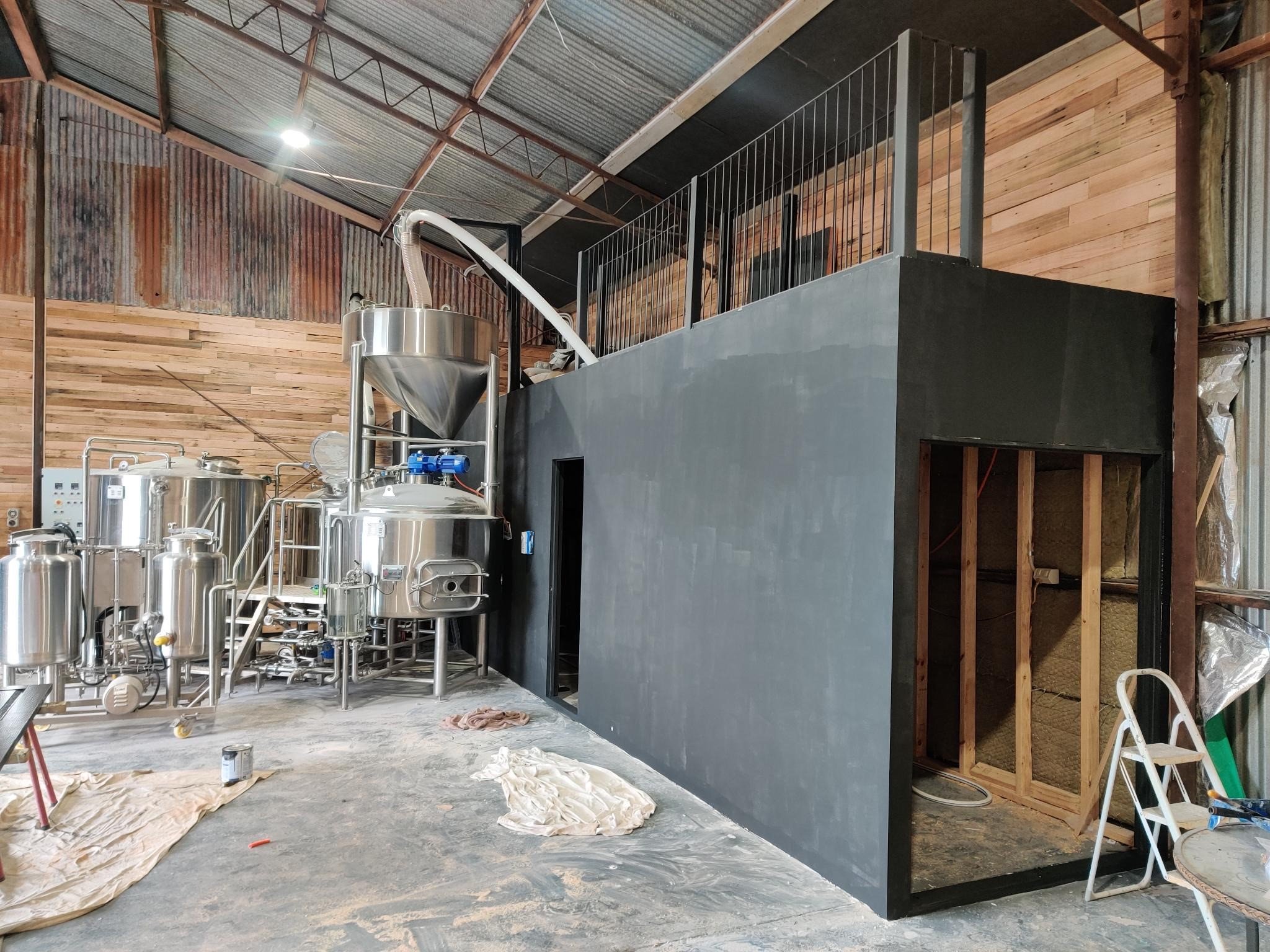 View inside a brewery or distillery with stainless steel brewing equipment and a black partially enclosed structure under construction, with wooden wall paneling and a ladder.