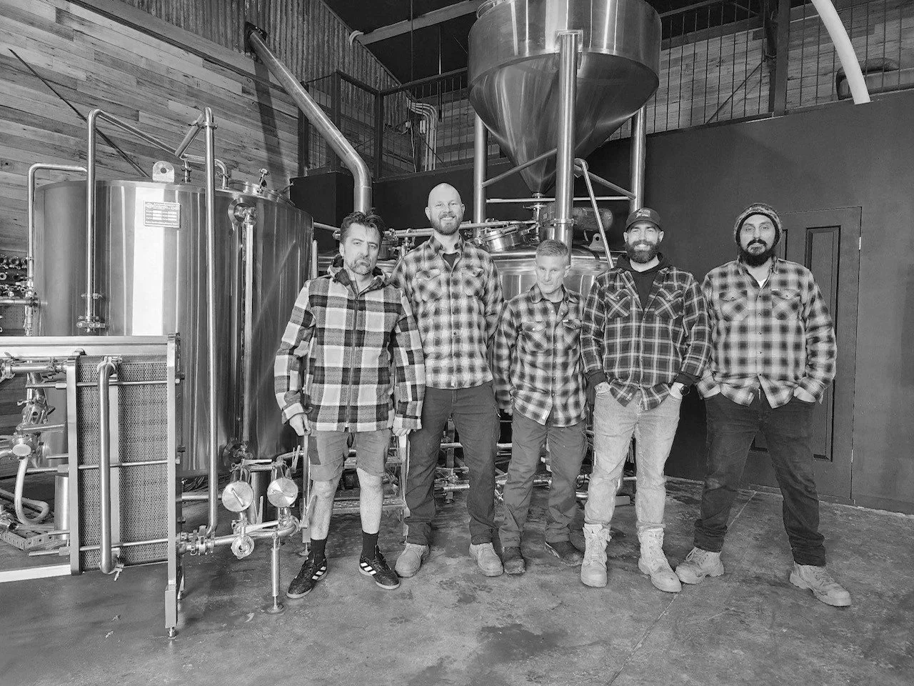 Five men wearing plaid shirts standing inside a brewery, with brewing equipment in the background.