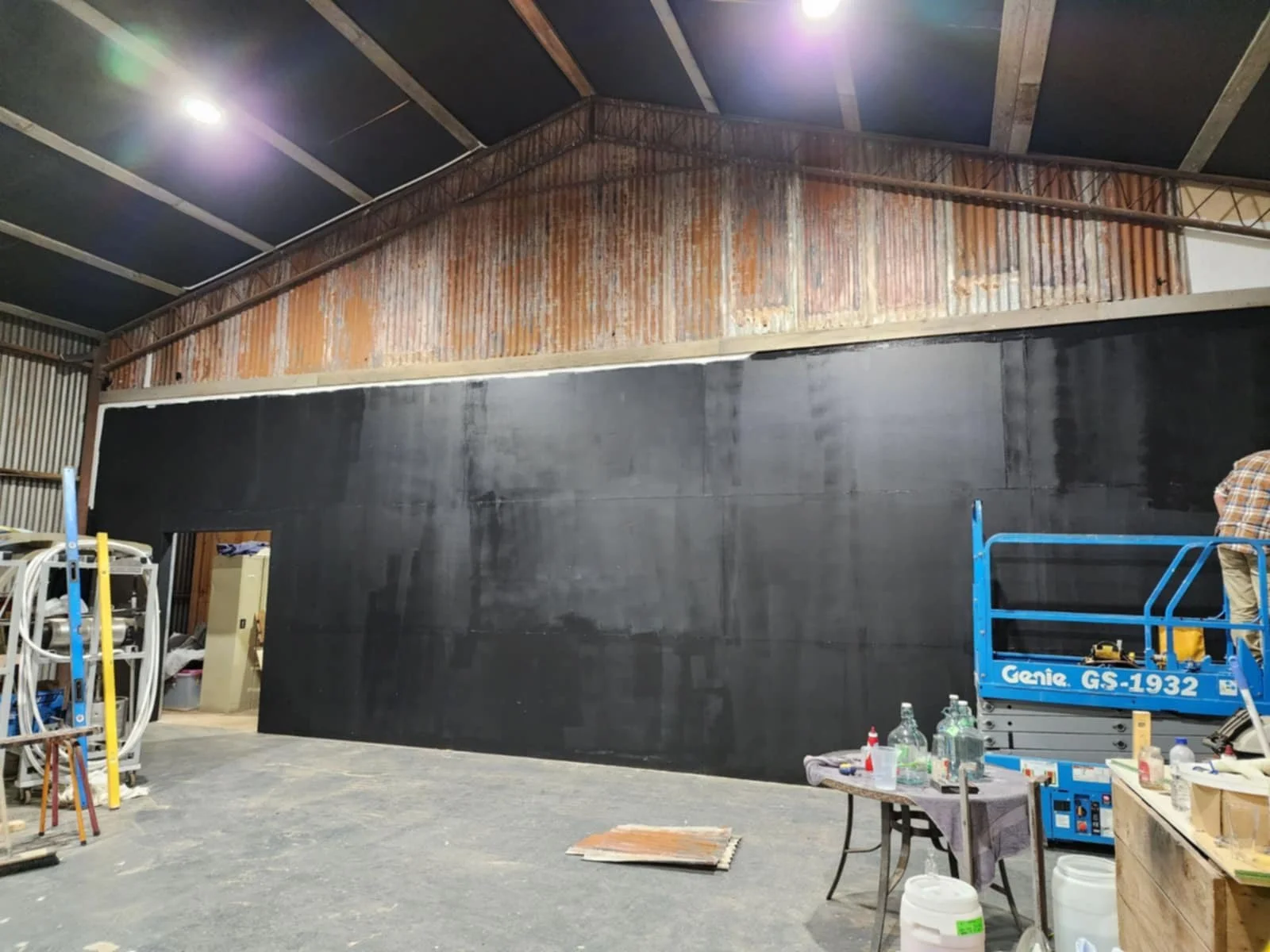 Interior of a warehouse or workshop with a large black chalkboard or painted wall, construction equipment including a blue scissor lift, work tables, and various bottles and tools on the tables.