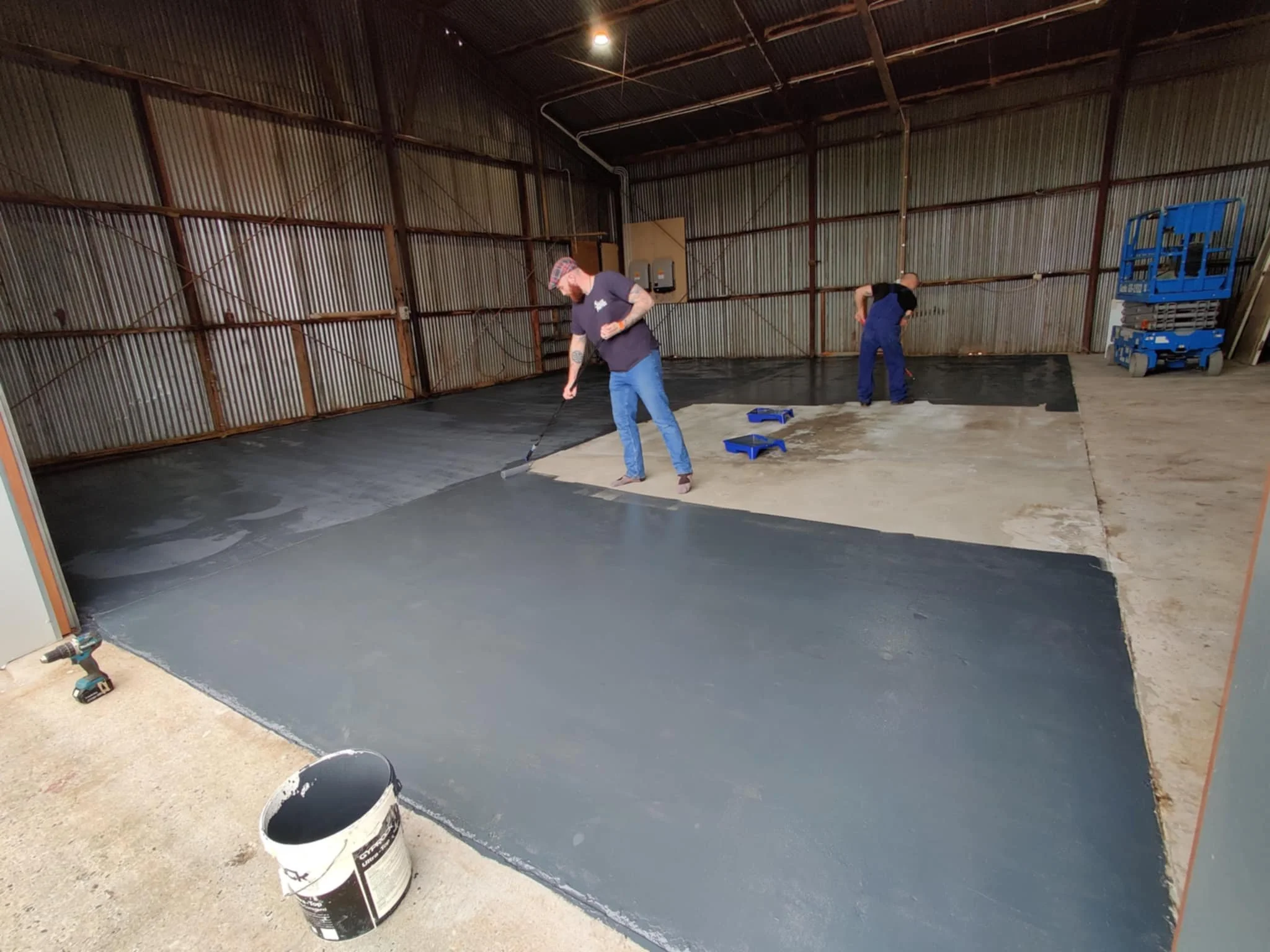 Two men are painting a concrete floor with black paint in a large, open industrial space with metal walls and ceiling.