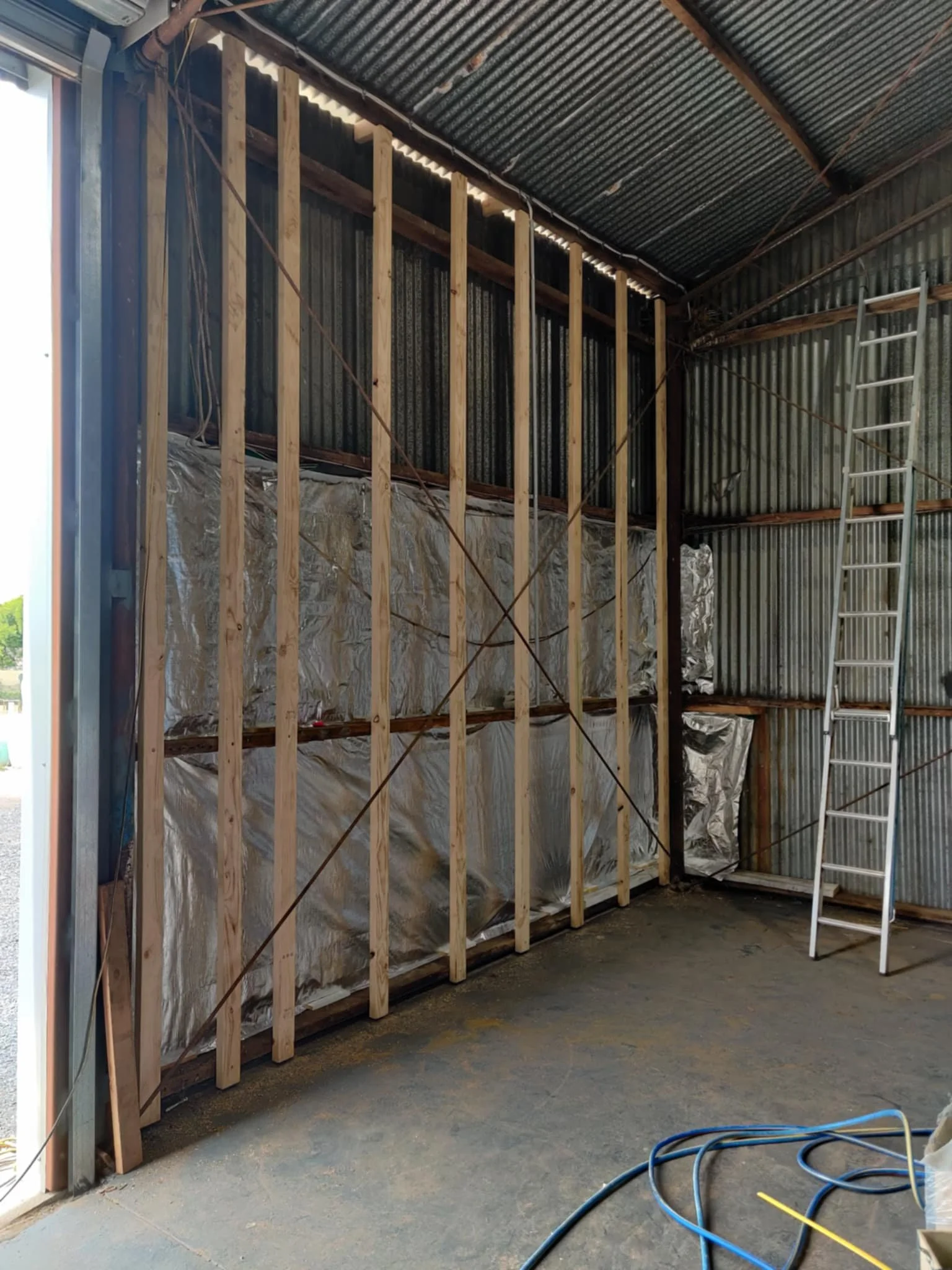 Interior of a garage under construction with exposed wooden framing, metal siding, a ladder, and construction cables.