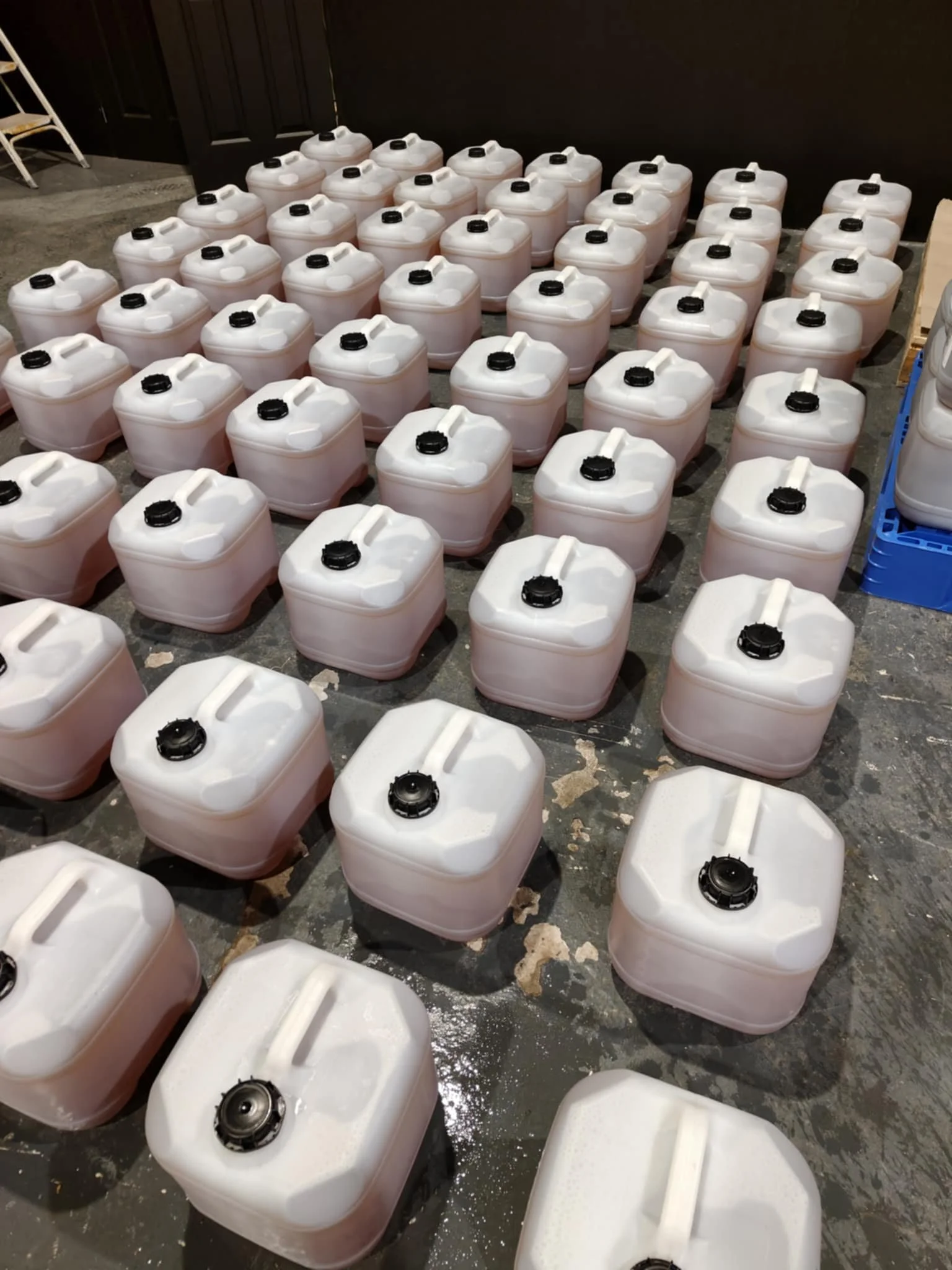 Numerous white plastic jerry cans with black caps placed on a gray concrete floor in a storage or warehouse setting.