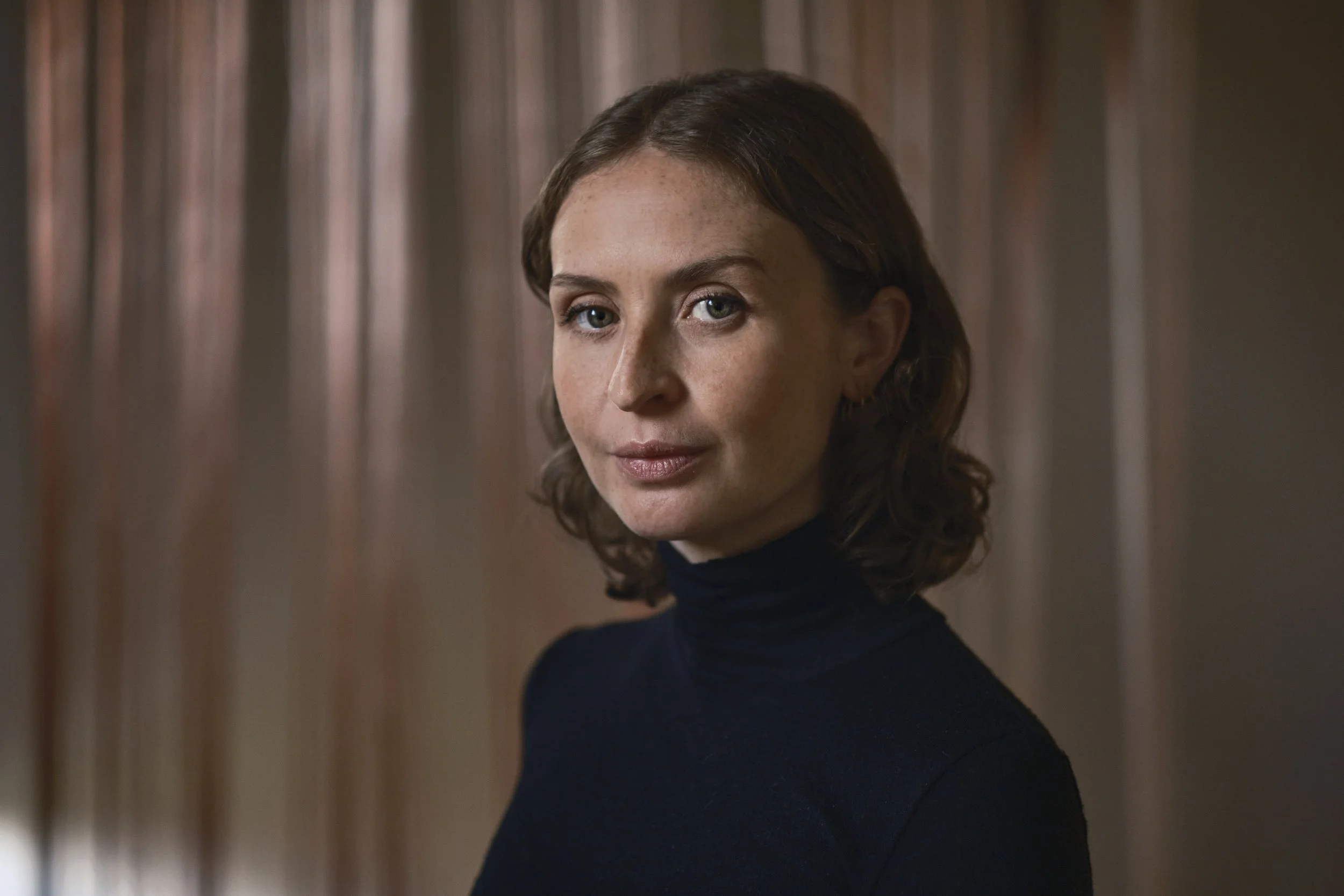 A woman with shoulder-length brown hair wearing a dark turtleneck, standing indoors against a background of vertical wood panels.