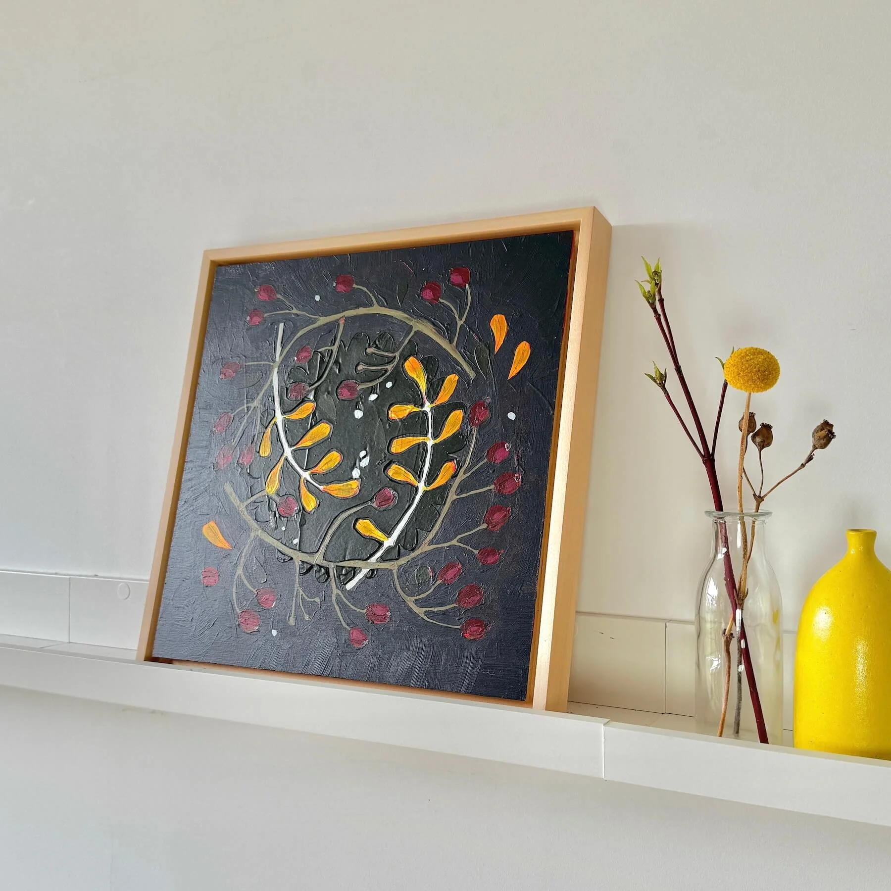 Angled left view of the framed painting Wilder by Ali Mackie Art on a white ledge alongside a yellow ceramic bottle and dried billy buttons.