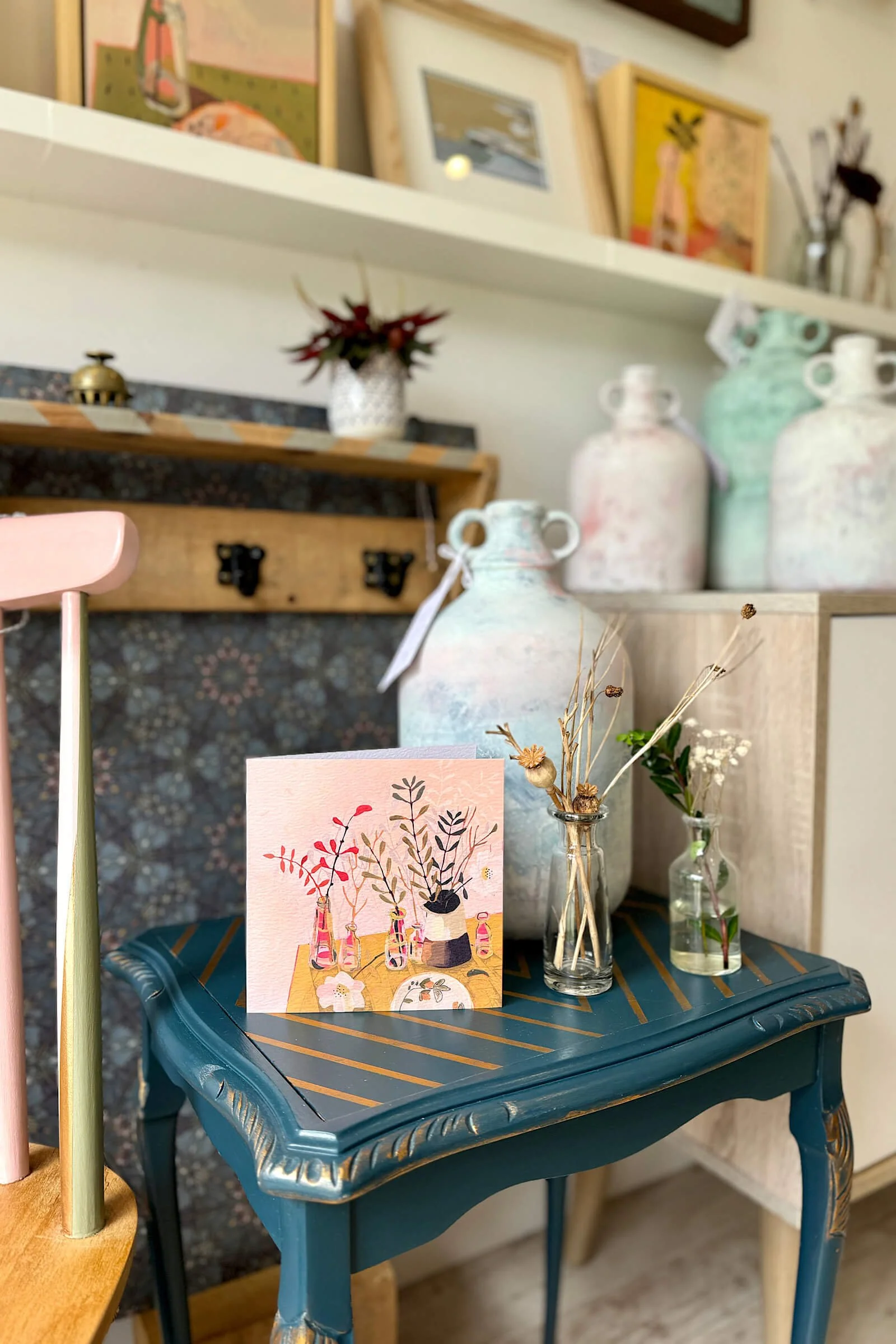 Decorative table with vases, dried flowers, and greeting card in a cozy interior setting.