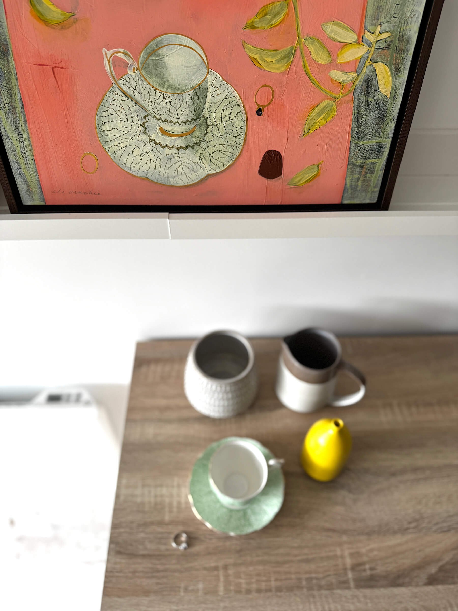 Top view of a wooden table with a small white plate on a green napkin, a yellow ceramic vase, two mugs, and a textured white cup. A painting hangs on the wall above the table, depicting a tea set, a cup, and some abstract elements on a pink backgroun