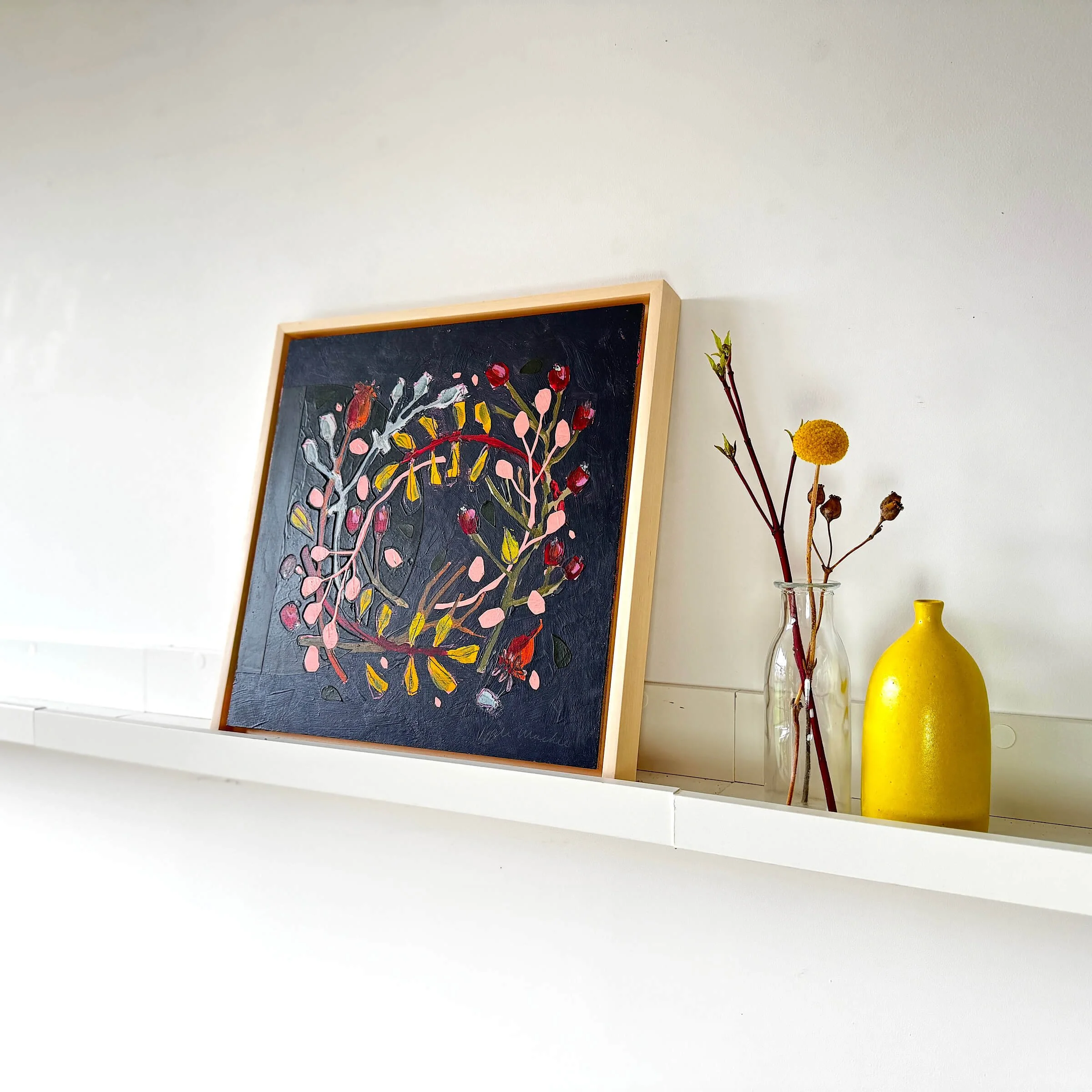 Wallis original mixed media botanical wreath painting by Ali Mackie displayed in a light wood tray frame on a white shelf. 