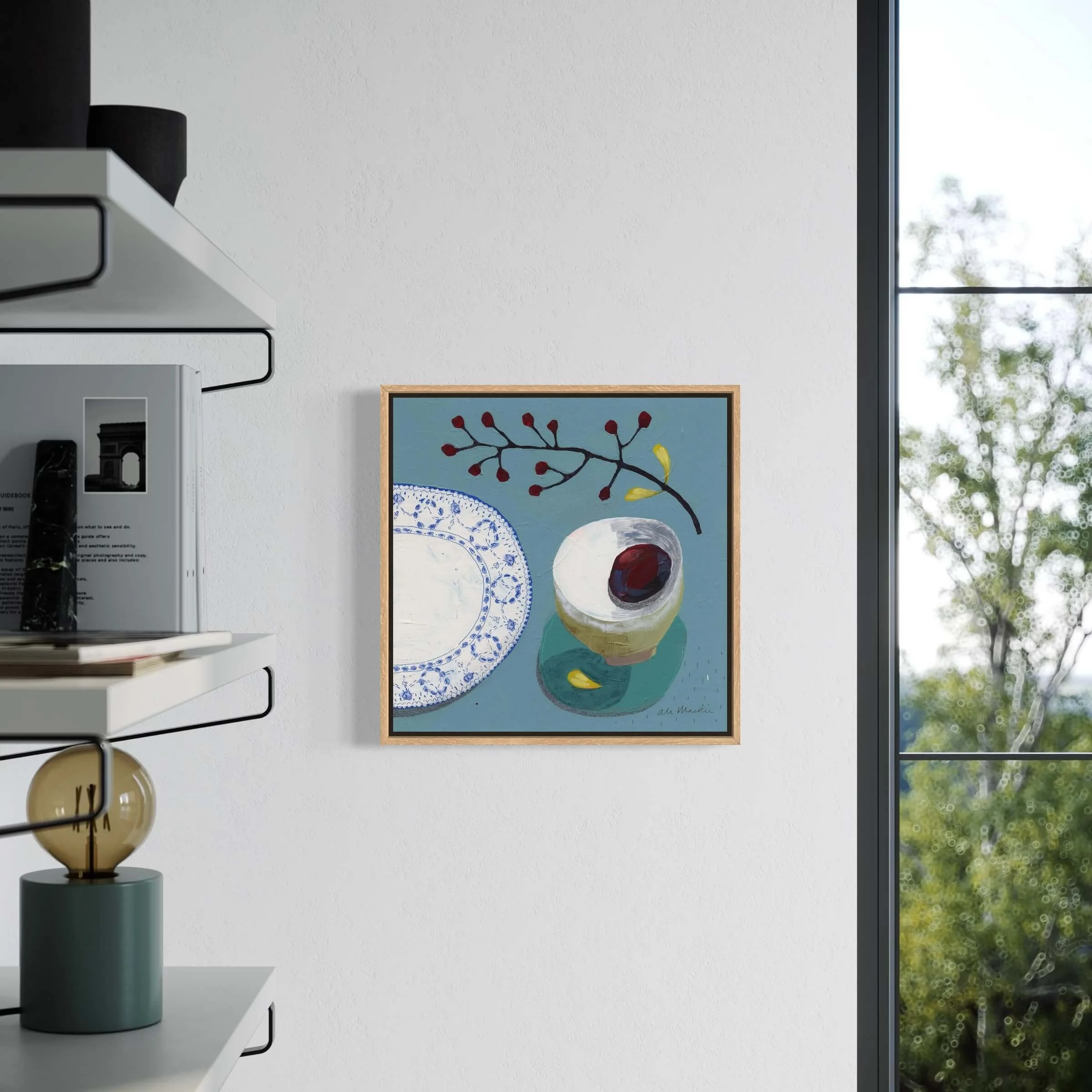 Bright living room interior mockup displaying framed blue still life artwork by Ali Mackie Art on a light wall near a window.
